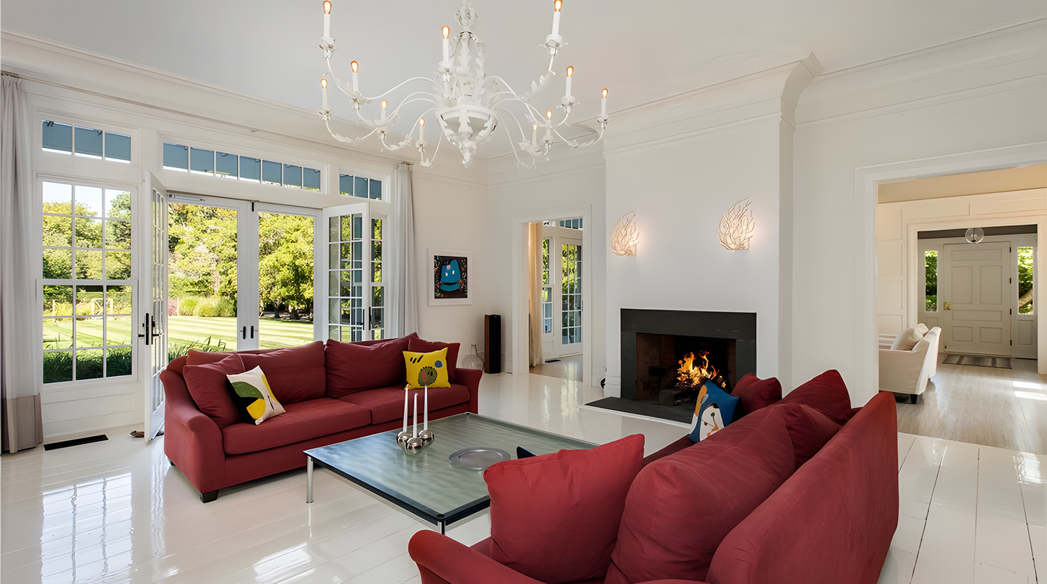 Bright living room with large windows, white walls, a white chandelier, and a fireplace with a fire. Red sofas with colorful pillows, a coffee table with candlesticks, and art on the wall.