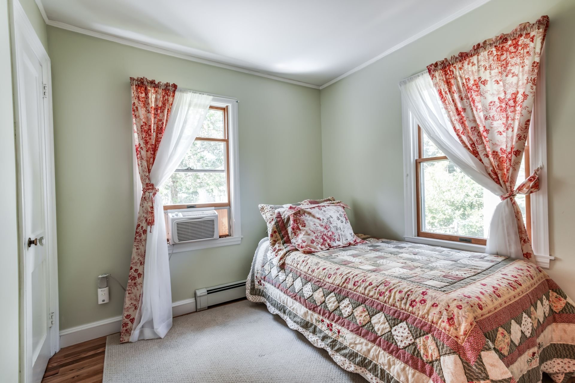 A bedroom with two windows dressed with floral curtains, a bed with a quilt and pillows, and an air conditioning unit in one window.