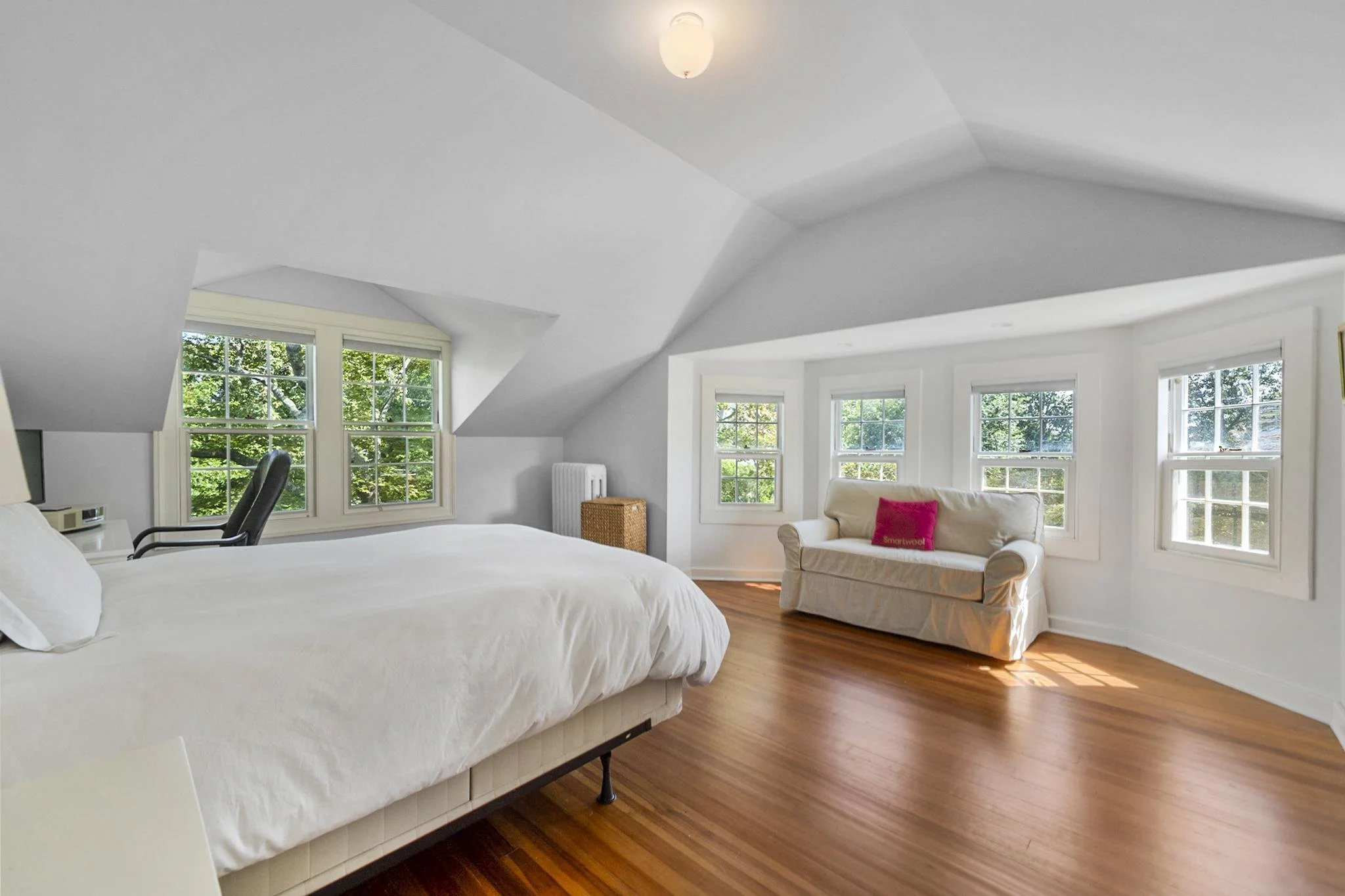A bright bedroom with white walls and sloped ceiling, six windows with greenery outside, a white bed, a black office chair, a beige sofa with a pink pillow, and hardwood floors.