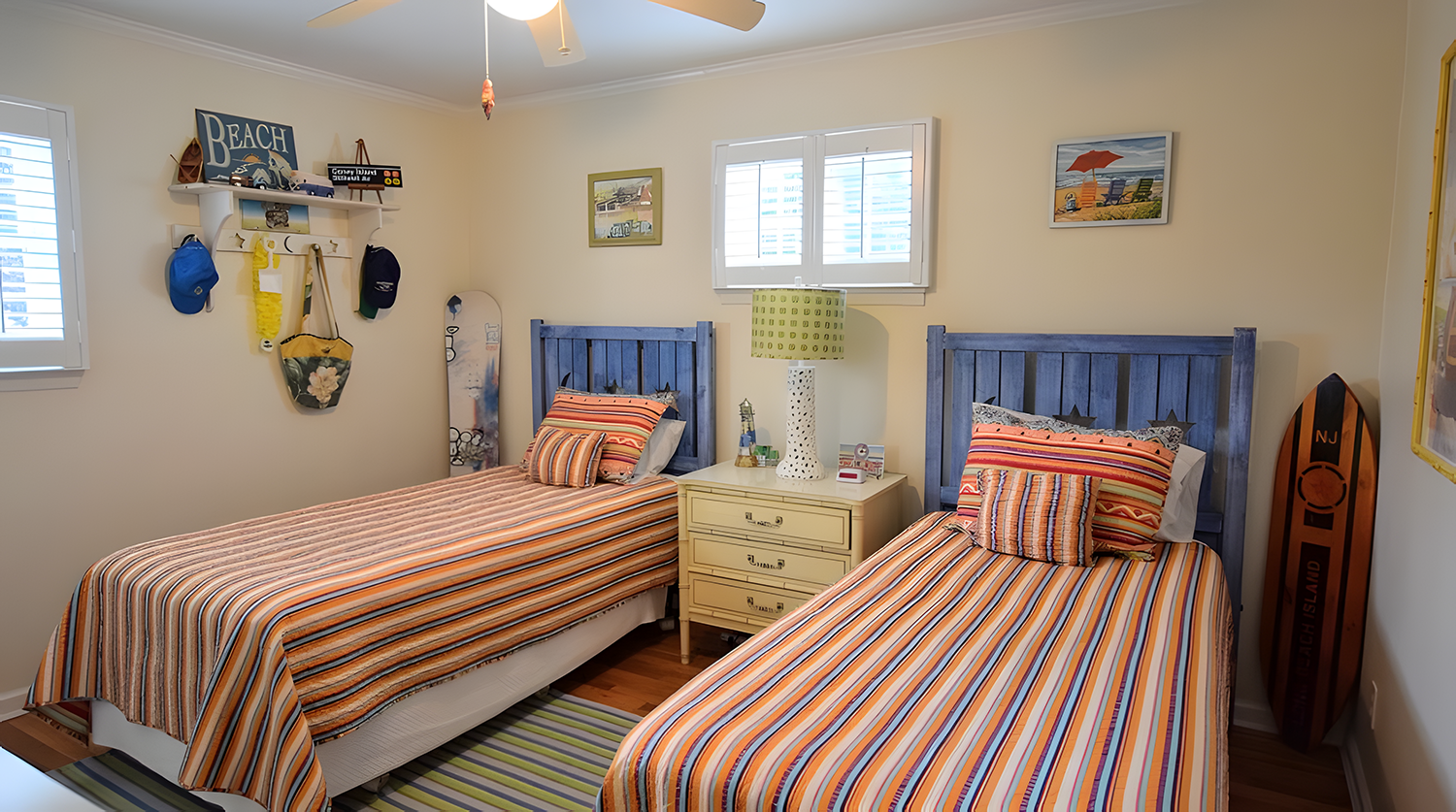 A bedroom with two twin beds with colorful striped bedding and matching pillows, a yellow nightstand with a lamp, small decor items, framed beach-themed pictures on the wall, and surfboards leaning against the wall.