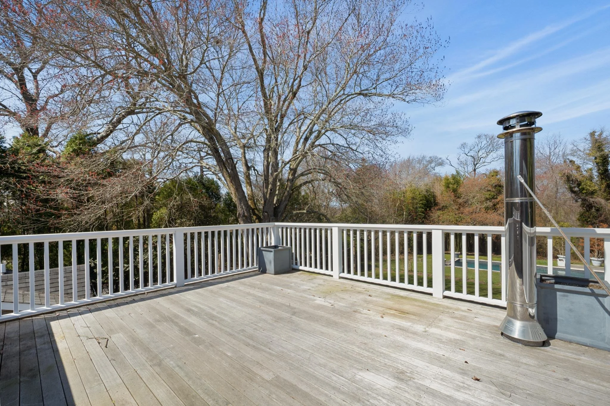 Empty wooden deck with white railing, outdoor heater, large tree with bare branches, and a swimming pool in the background, on a clear day.