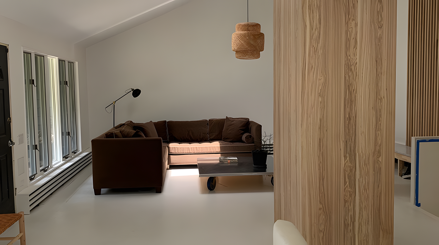 Minimalist living room with a brown sofa, black standing lamp, wooden panel wall, light-colored floor, and a small coffee table with magazines, beside a black door and windows.