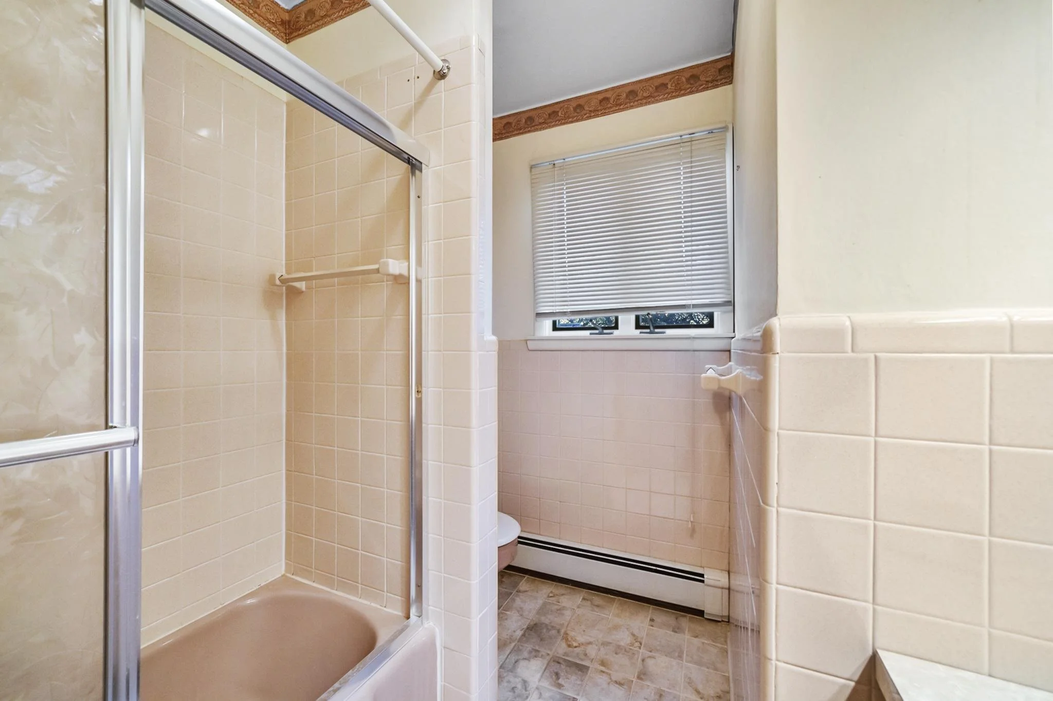 A bathroom with a walk-in shower, beige tiled walls, and a window with closed blinds.