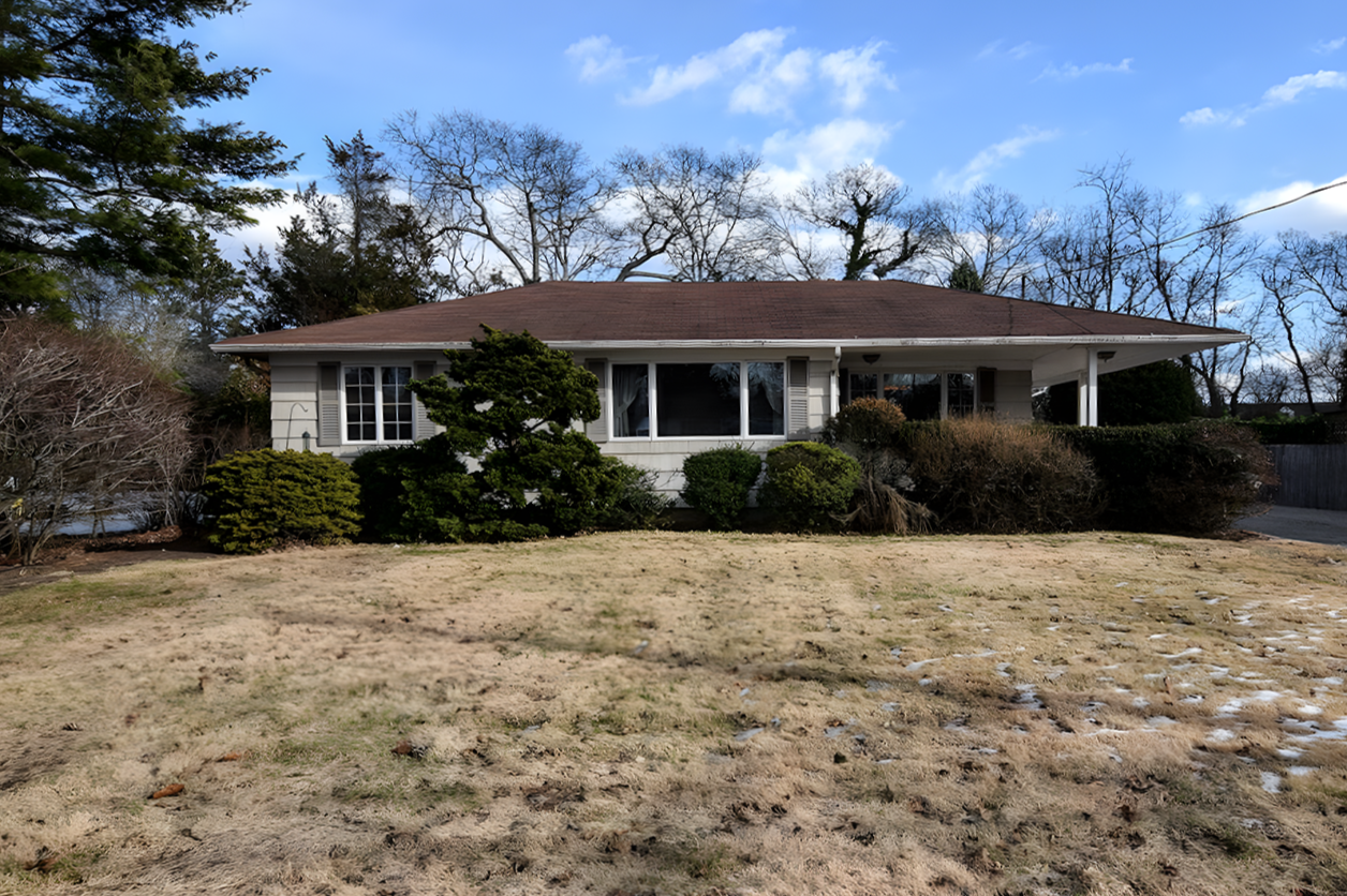 A single-story house with a brown roof, white exterior, and large front windows, surrounded by bushes and trees, with a front yard that has patches of snow and dry grass.