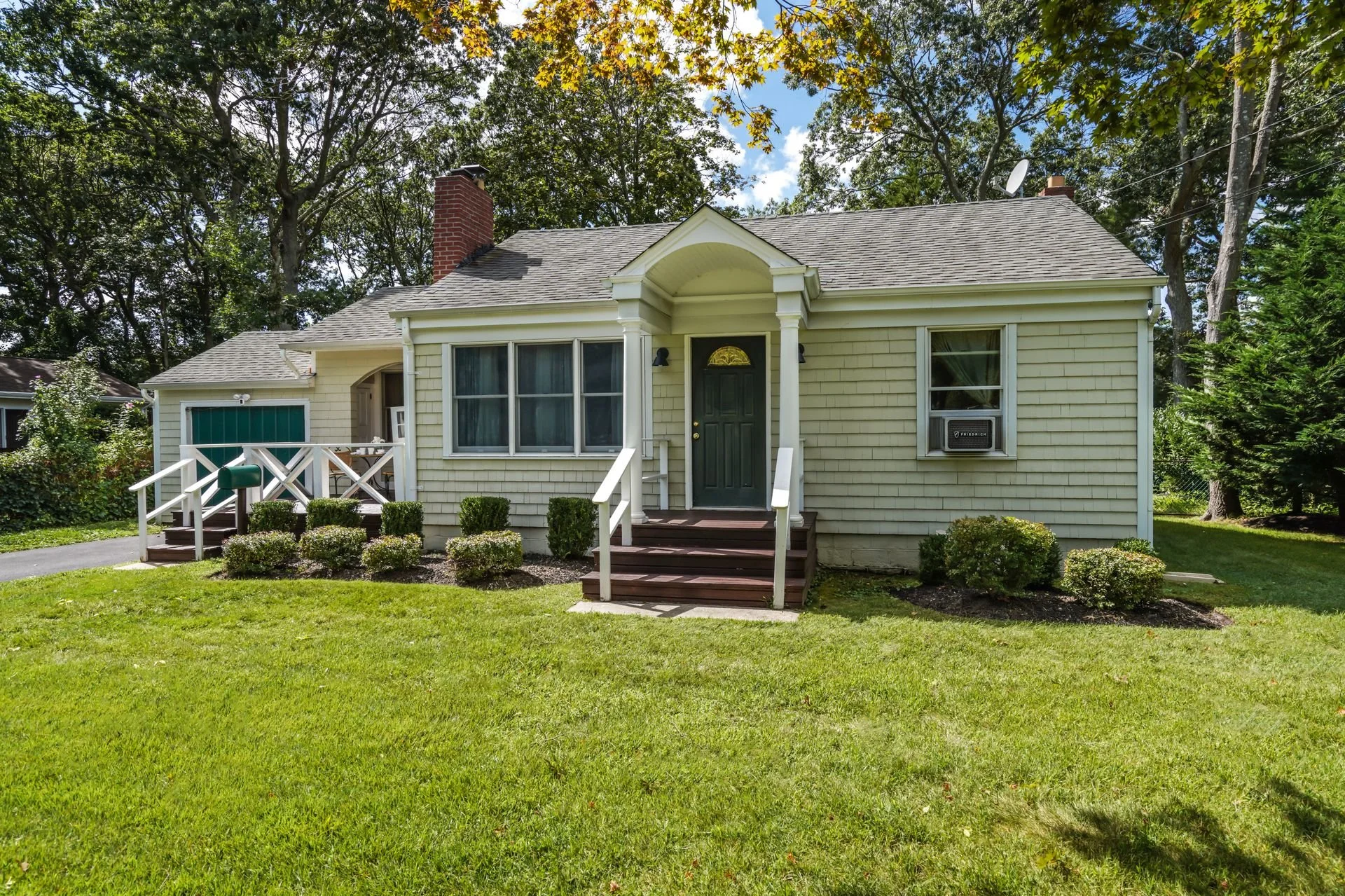 Front view of a small yellow house with a front porch, surrounded by green grass and bushes, trees in the background, and a clear blue sky.
