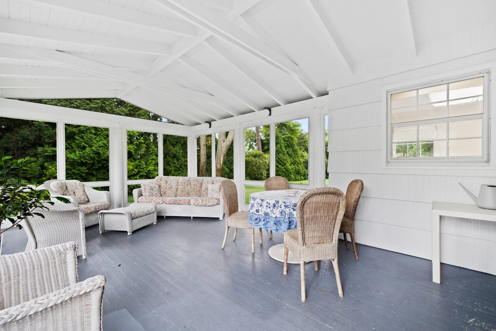 Enclosed porch with white wicker and fabric seating, wicker chairs, a round table with a blue floral tablecloth, and large windows revealing green trees outside.