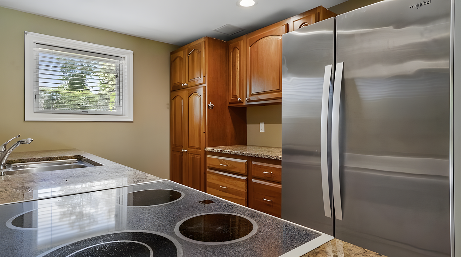 Modern kitchen with wooden cabinets, granite countertops, stainless steel refrigerator, and a window with blinds overlooking trees.
