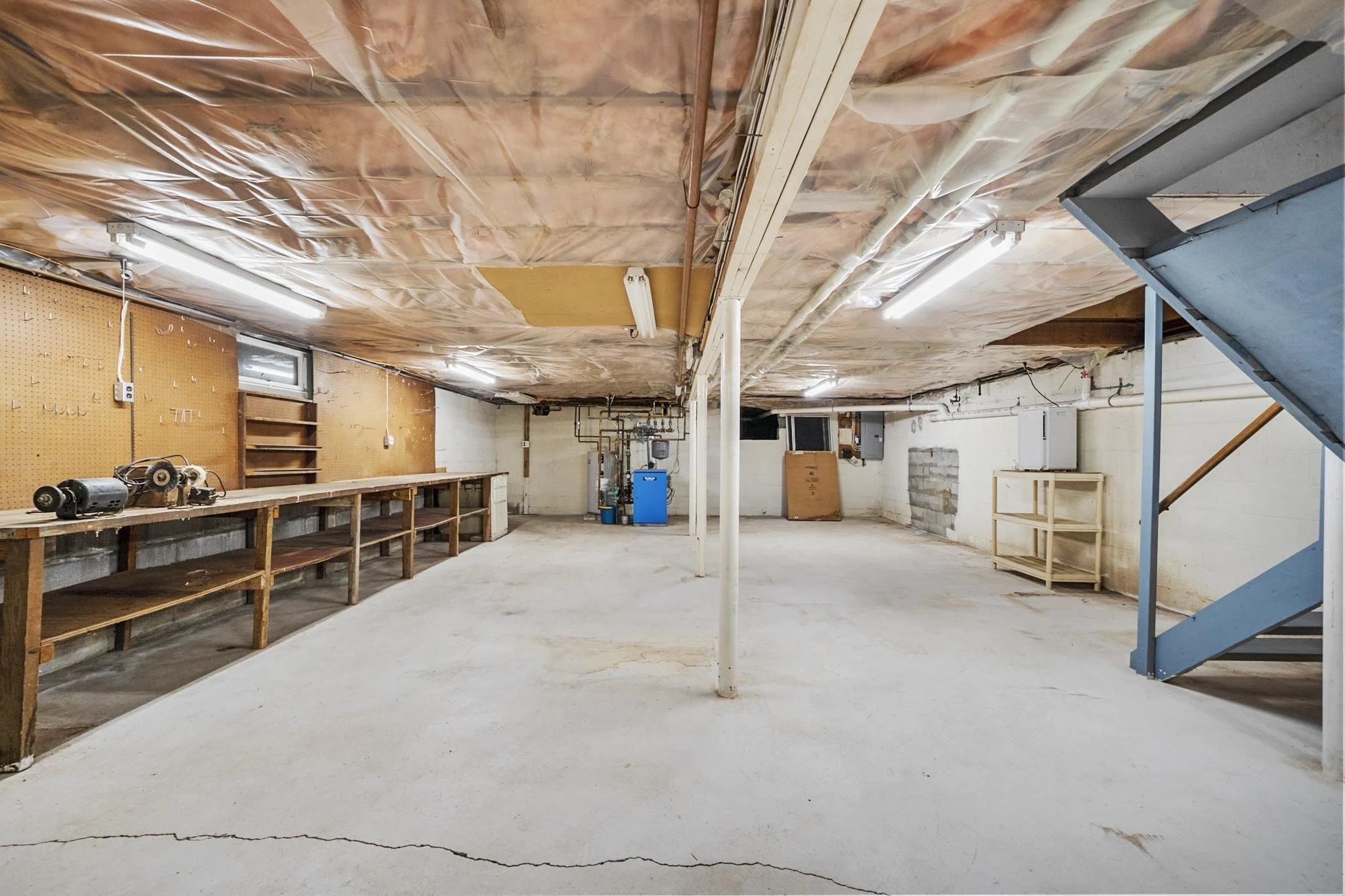Empty basement or garage with a concrete floor, unfinished ceiling with exposed insulation and pipes, a workbench along one wall, a blue water heater or furnace, and some shelving and equipment.