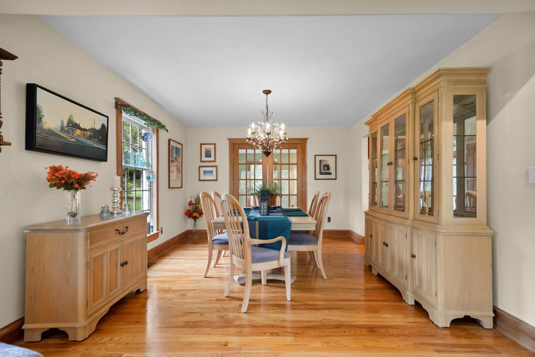 A dining room with a wooden dining table, chairs, a large wooden china cabinet, and a chandelier, with framed pictures on the walls and windows with greenery outside.