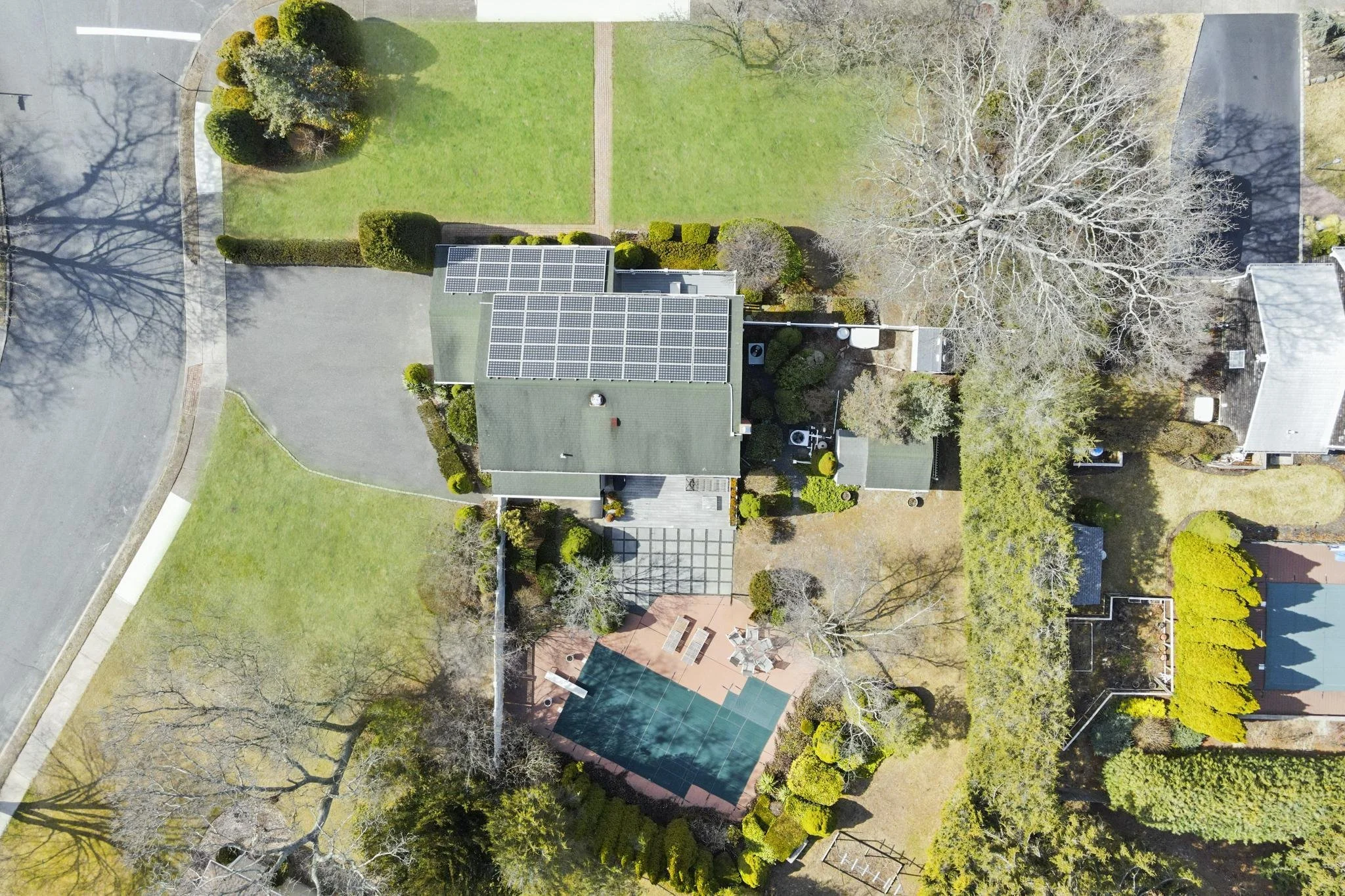 An aerial view of a house with solar panels on the roof, surrounded by trees, lawn, and neighboring houses, with some trees bare and others with green foliage.