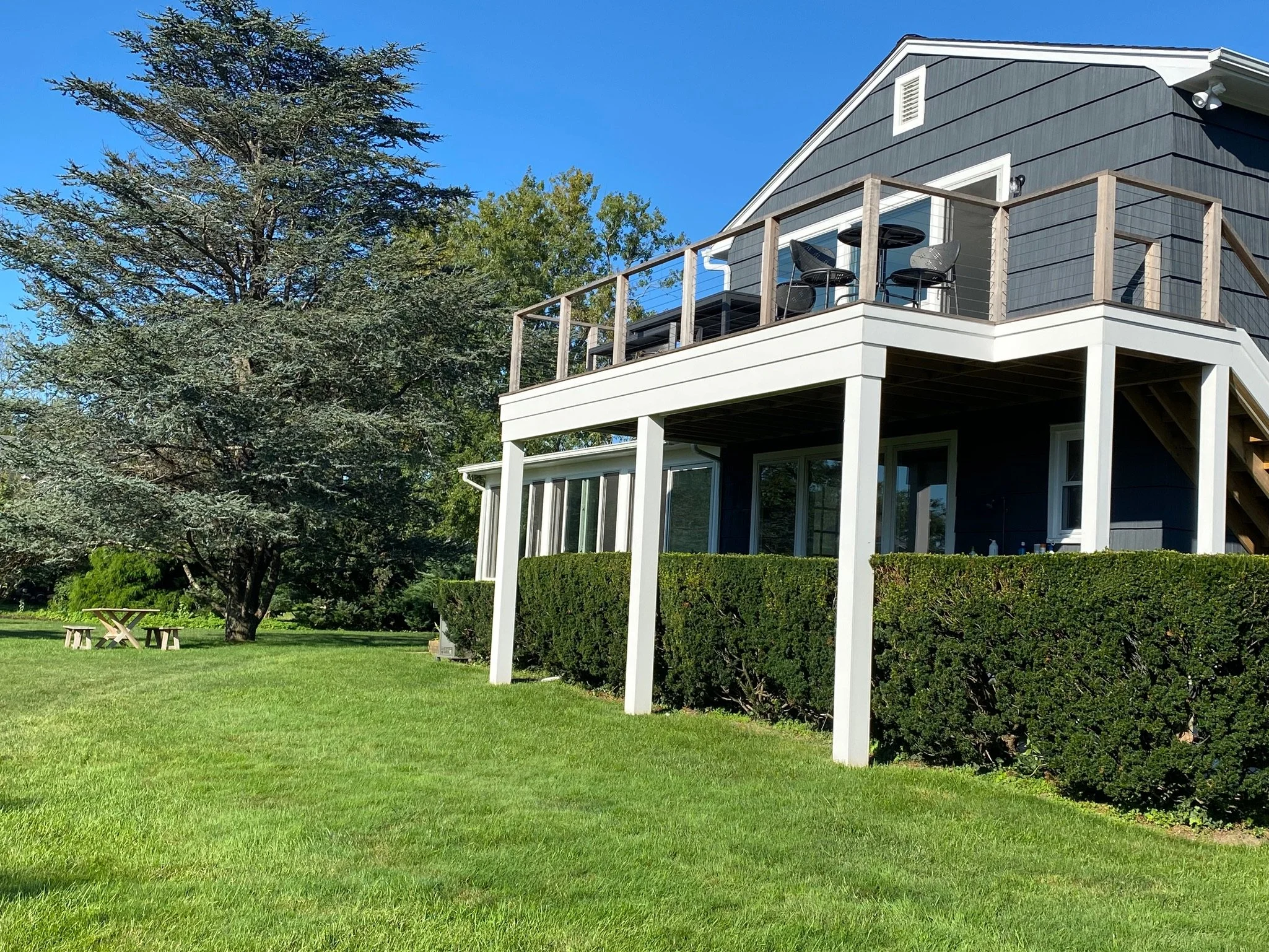 A house with a second-story balcony, black siding, and white trim, surrounded by green trees and a well-maintained lawn, with a picnic table in the yard.
