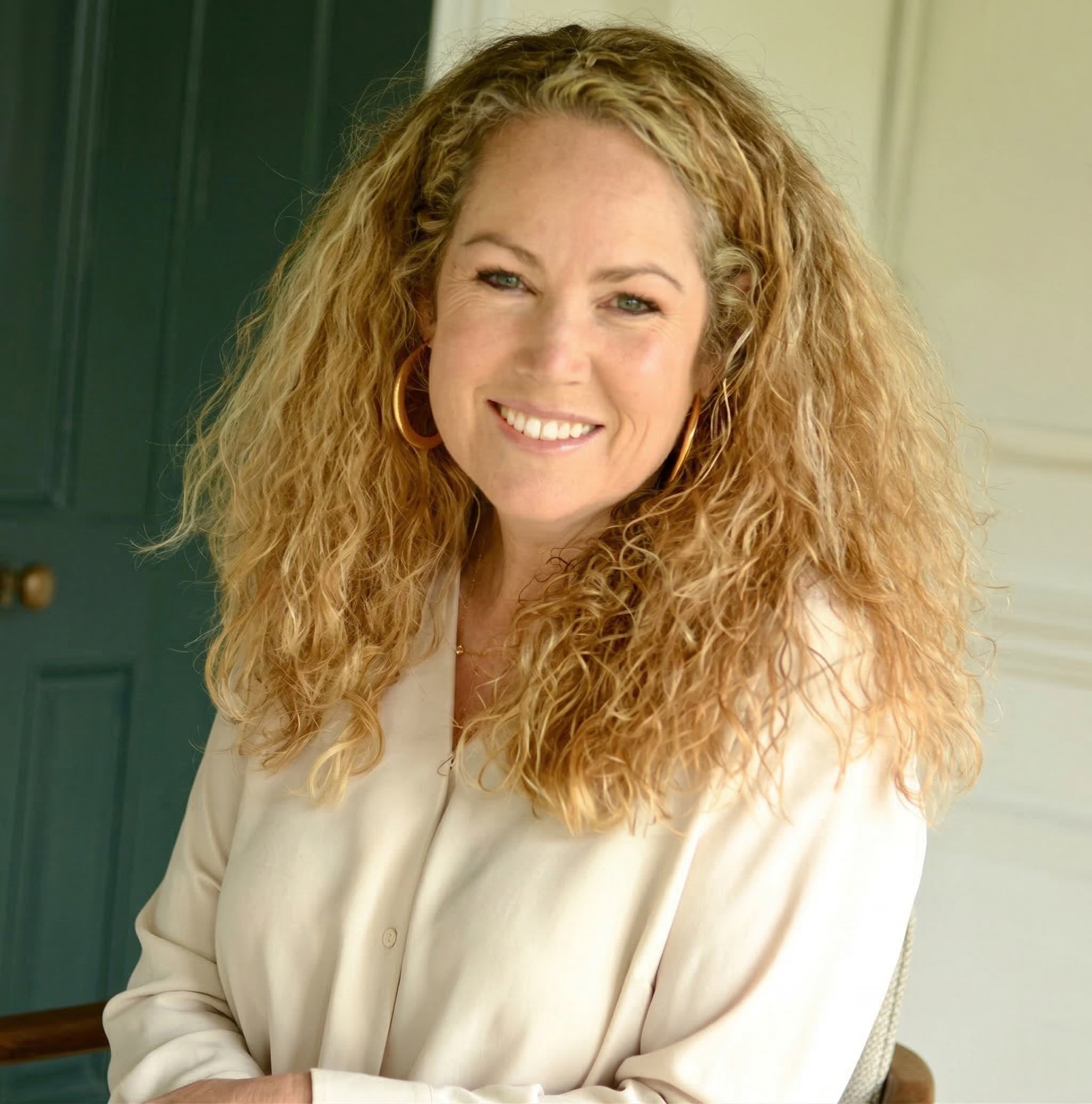 A woman with curly blonde hair, wearing a white blazer and gold hoop earrings, is smiling. She is sitting at a desk or table, with part of her arm visible in the foreground. The background features a green door and a light-colored wall.