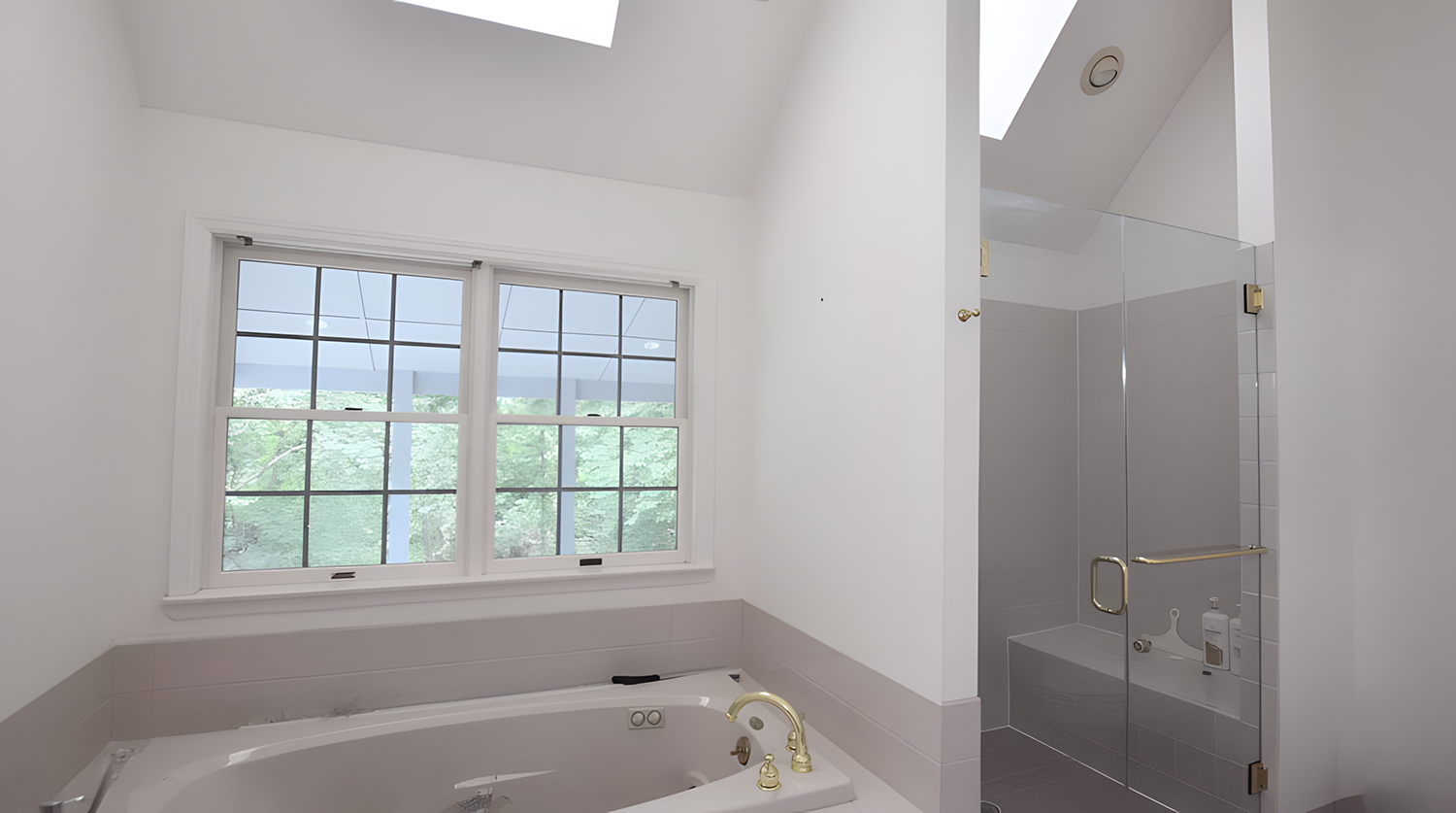 A bathroom with a large window, a corner bathtub with gold fixtures, and a glass-enclosed shower with a built-in shelf and a gold handle.