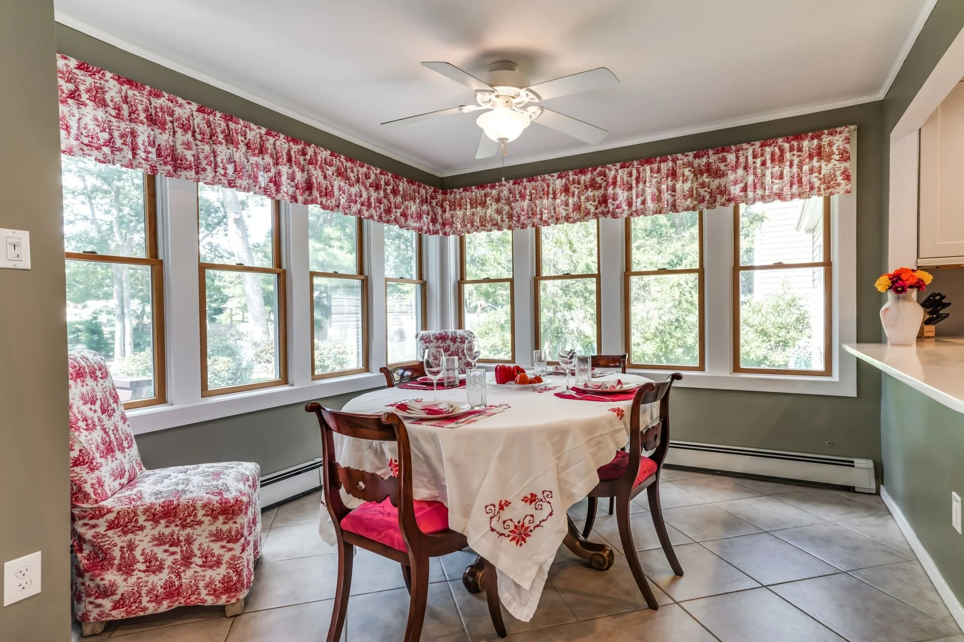 A dining room with a round table set for a meal, surrounded by wooden chairs with red cushioned seats. The table is covered with a white tablecloth decorated with red embroidery, and has glassware, napkins, and a bowl of orange and red orbs. The room