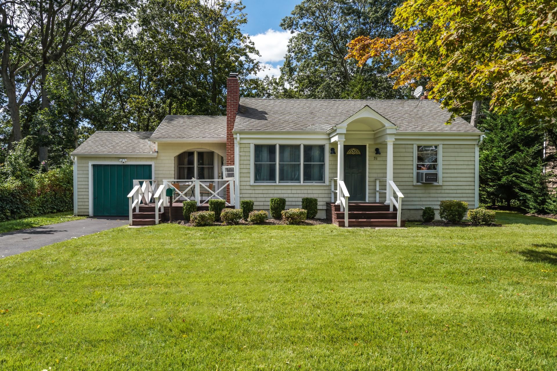 A yellow house with a front porch, green door, driveway on the left side, surrounded by green lawn and trees, with a chimney and satellite dish on the roof.