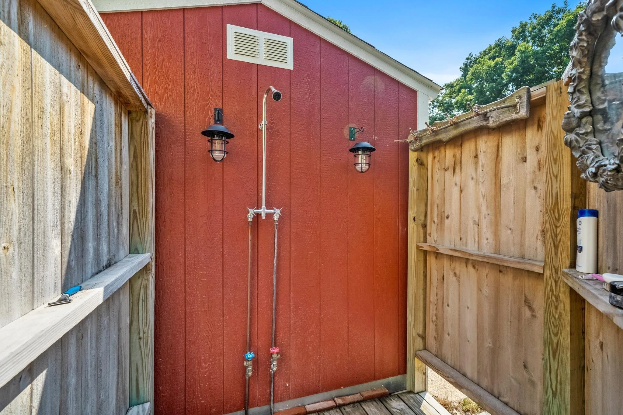 Backyard outdoor shower with red house wall, wooden fencing, and blue sky.