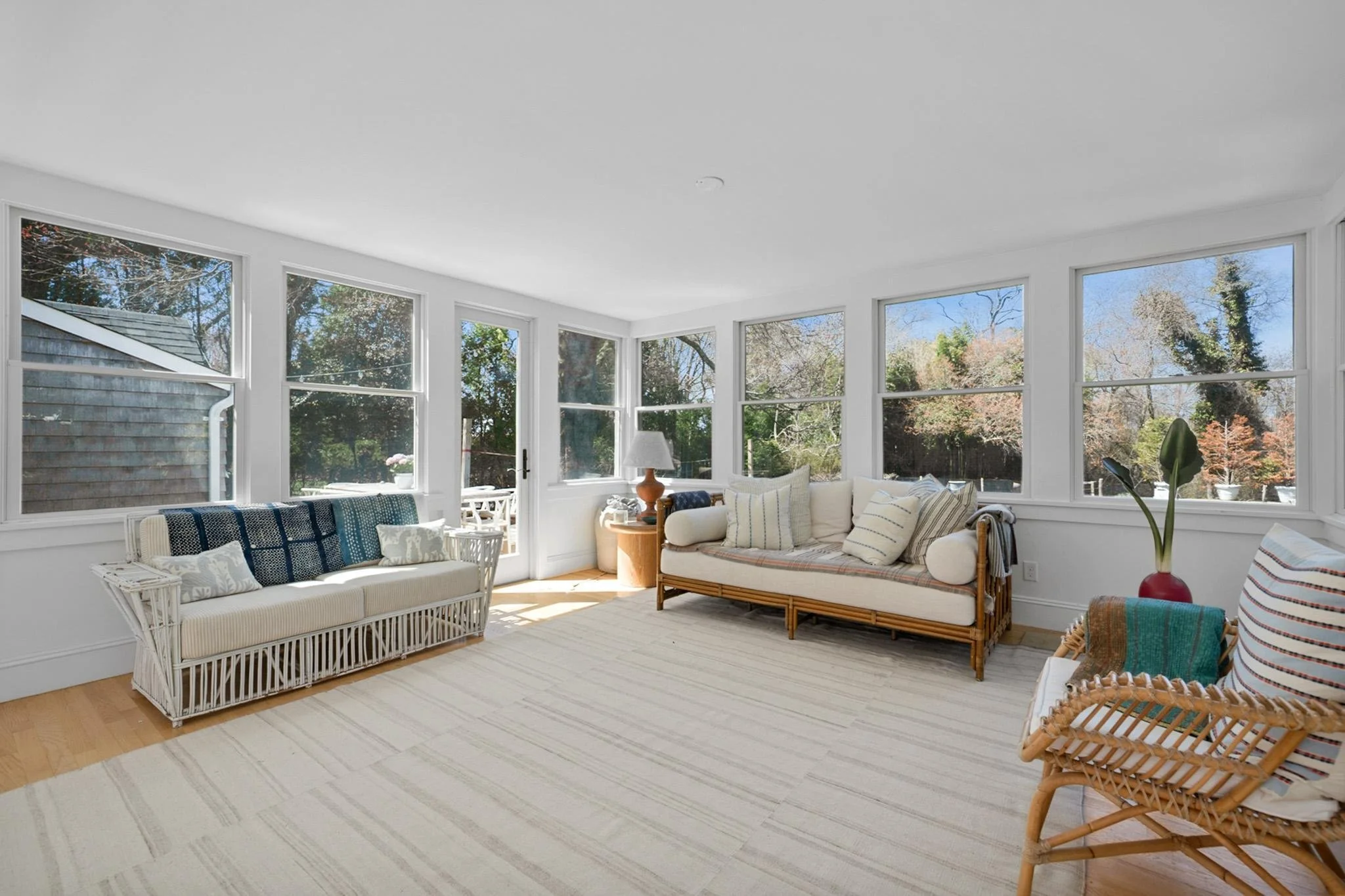 Bright sunlit living room with large windows showing trees outside. Contains white wicker and wooden furniture with cushions and blankets, a table with a lamp, and a rug on a wooden floor.