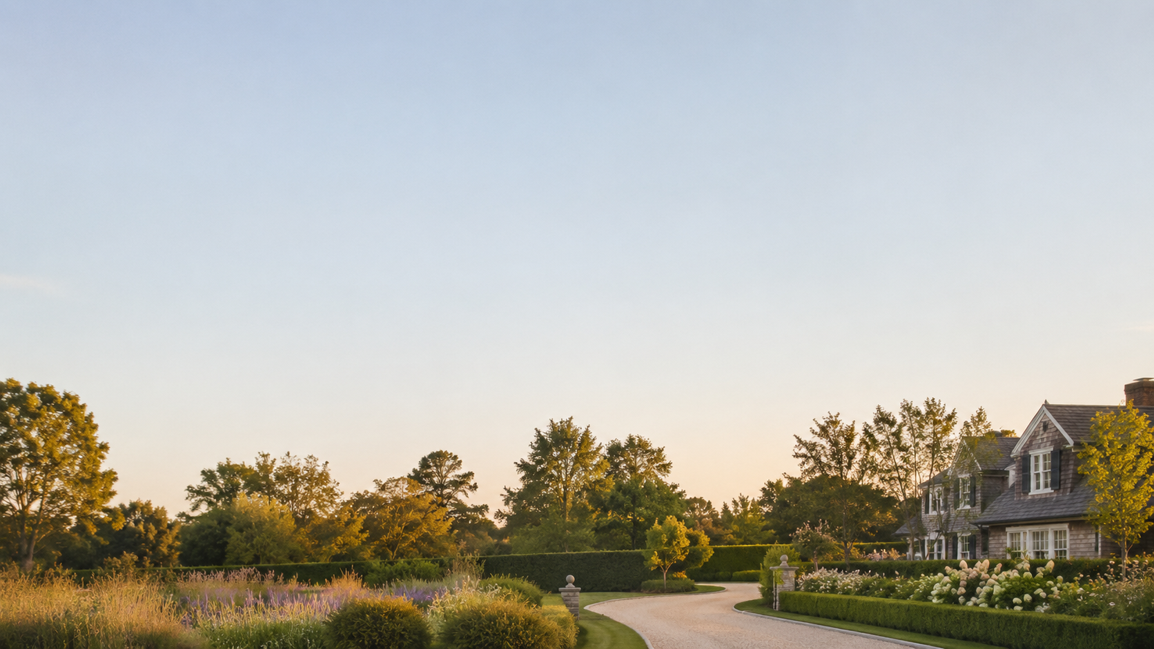 A large house with a landscaped front yard and a winding driveway under a clear sky at sunset.