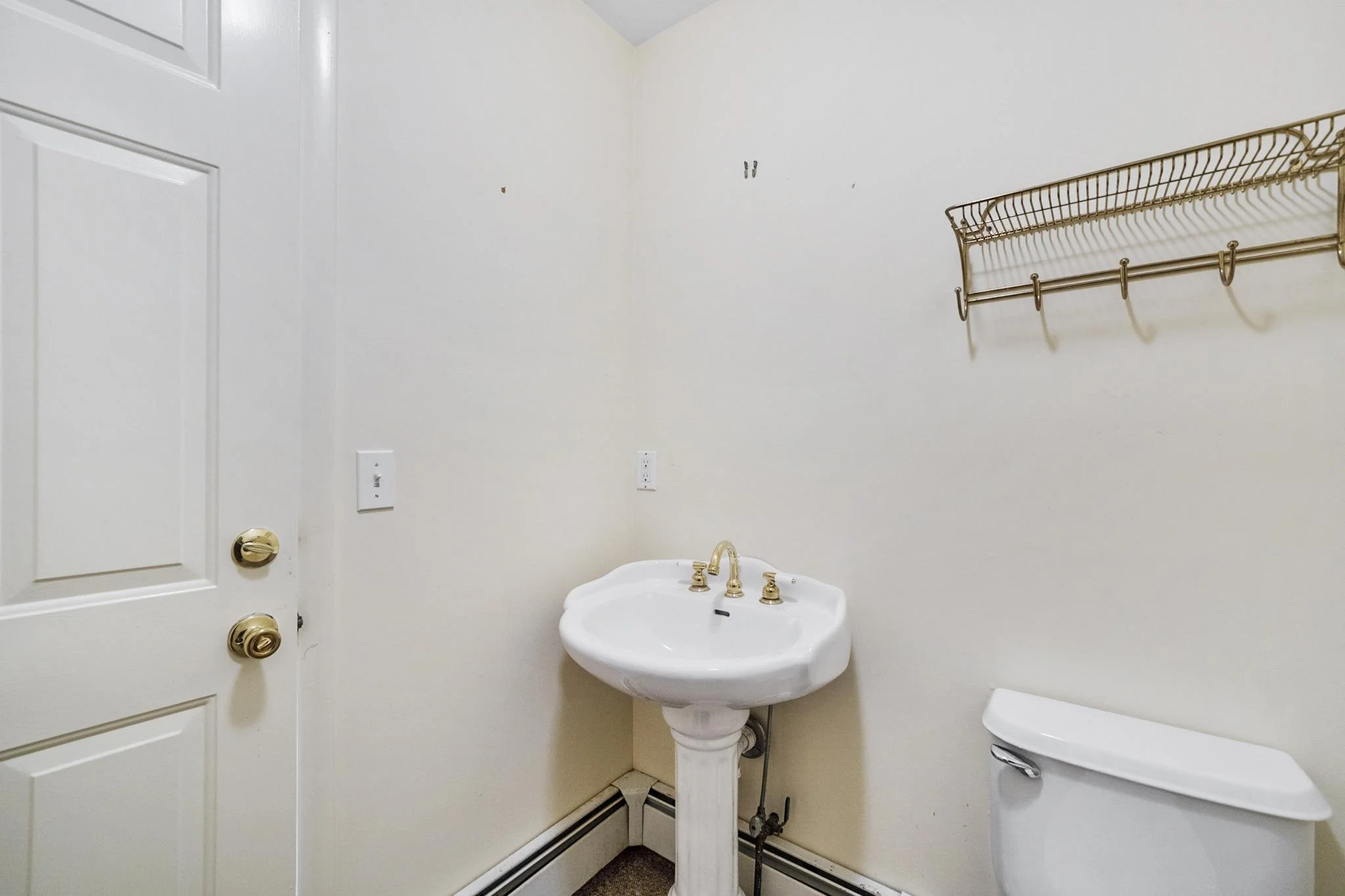 Small bathroom with a white pedestal sink, a toilet, a brass towel rack, and a white door with brass doorknob and lock. The bathroom has plain beige walls and baseboard heater.