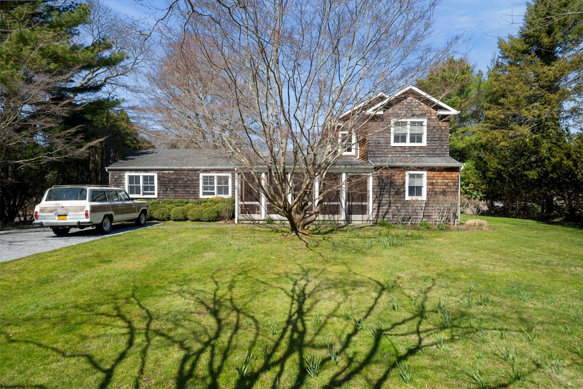 A house with weathered wooden shingles on the exterior, surrounded by a green lawn and bushes, with a leafless tree in front and other trees with green foliage in the background. A vintage station wagon is parked on a driveway to the left of the hous