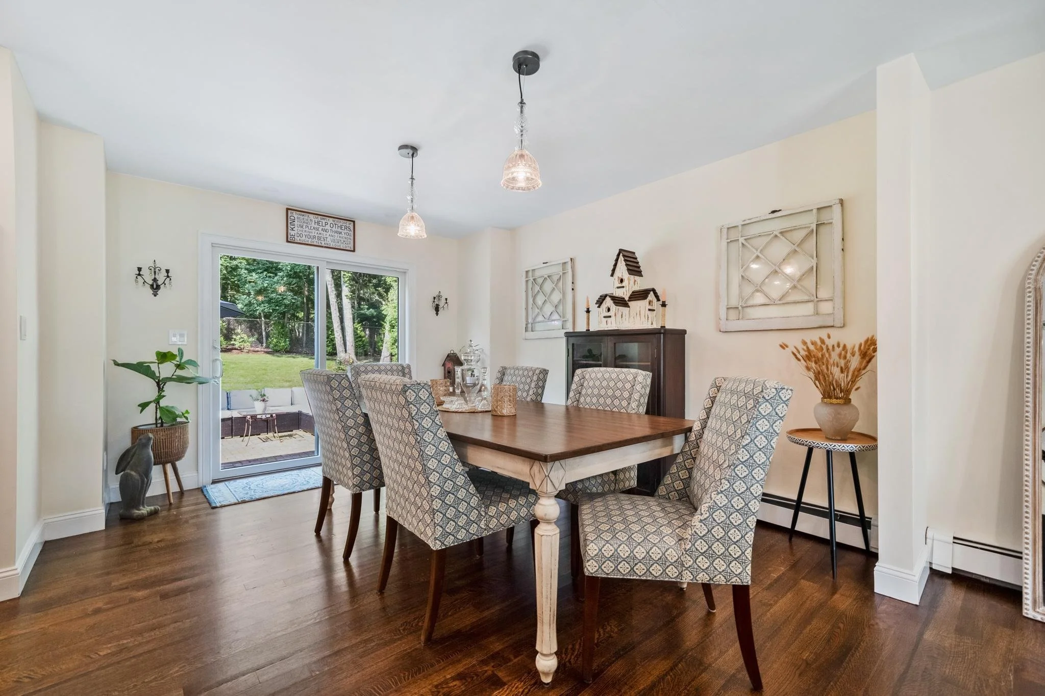 A dining room with a wooden table surrounded by patterned chairs, a sliding glass door leading to a backyard patio, and decorative items including a potted plant, a small cabinet with a dollhouse, and wall art.