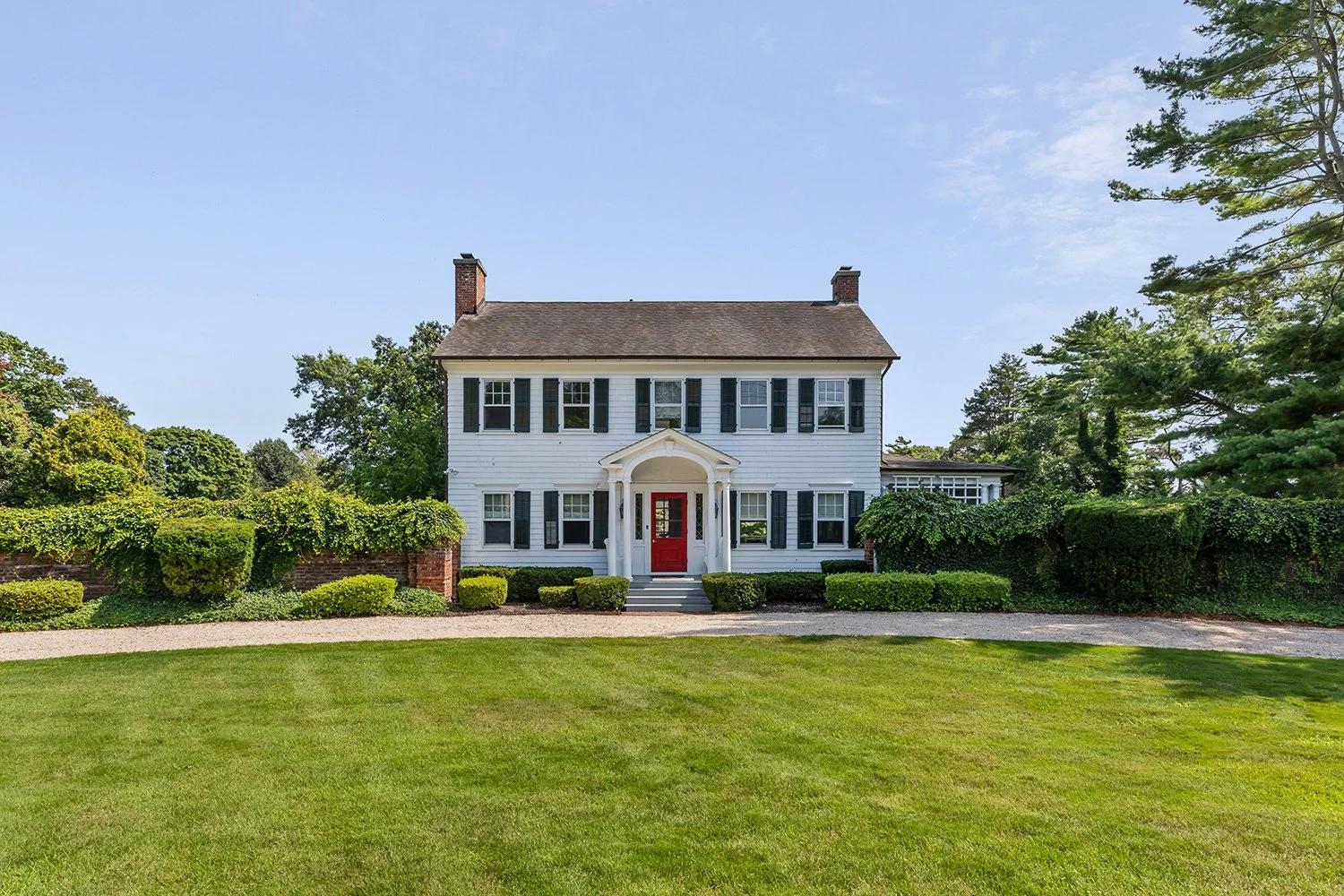 A large white porch-front house with black shutters, a red front door, and a well-kept green lawn.