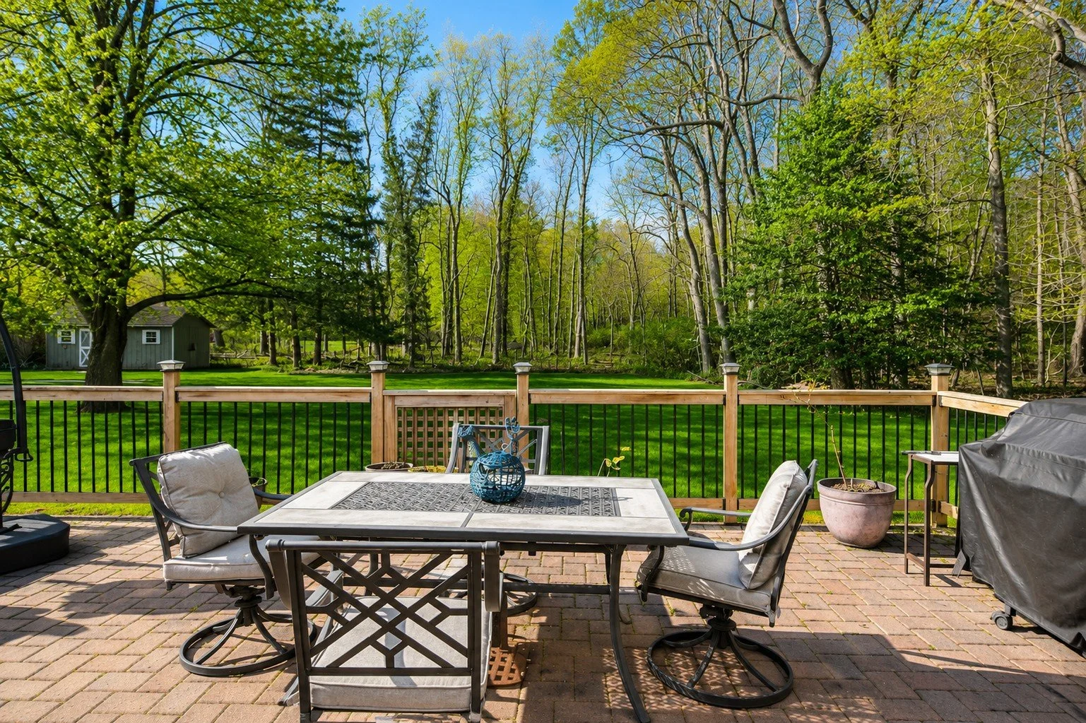 A backyard patio with a glass dining table and four cushioned chairs, a grill covered with a black protective cover, and potted plants, overlooking a green lawn with trees and blue sky.