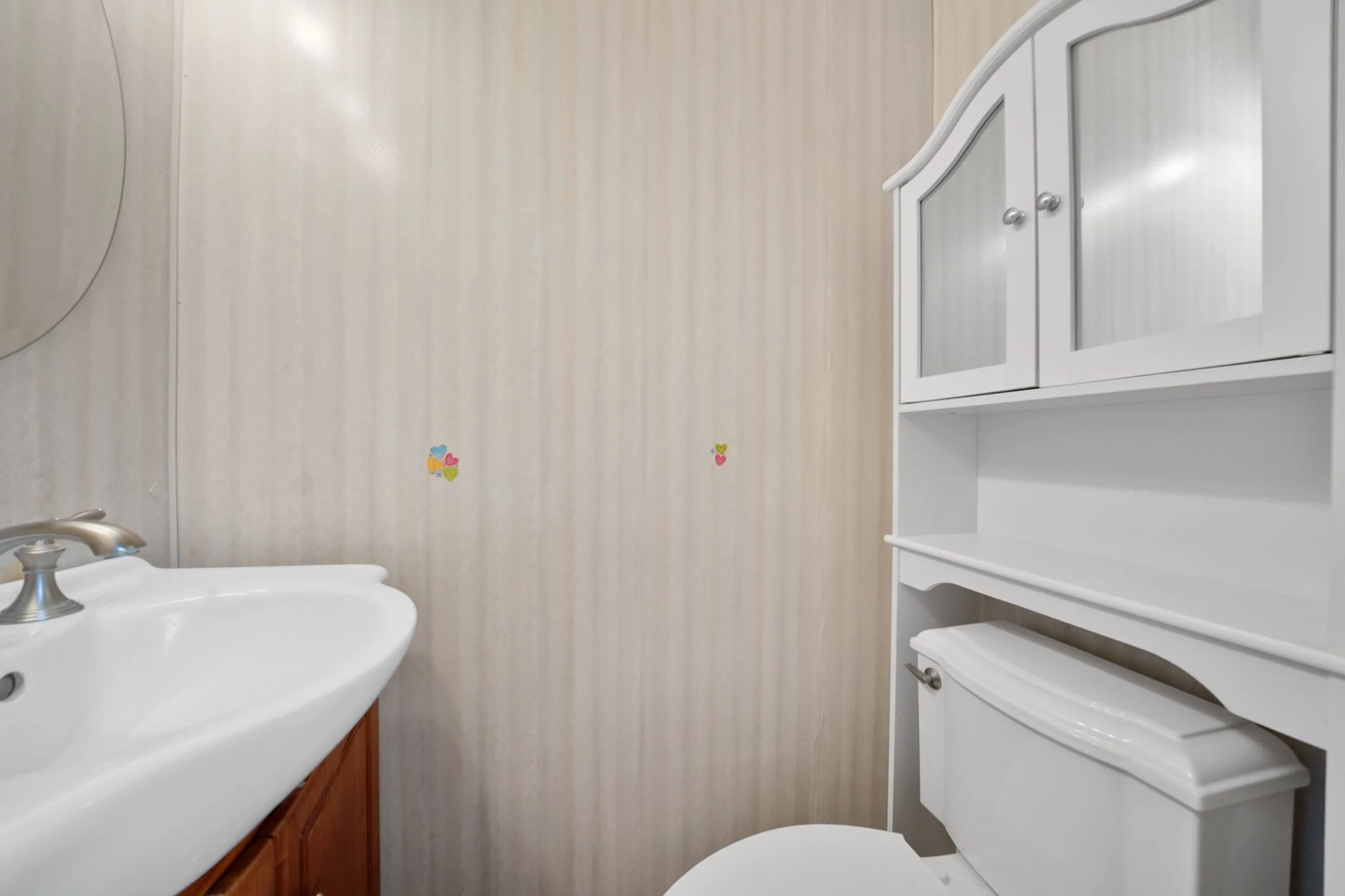 Bathroom with light-colored striped wallpaper, a white cabinet with glass doors, a white toilet, and a sink with a metal faucet. Small colorful heart stickers are on the wall.