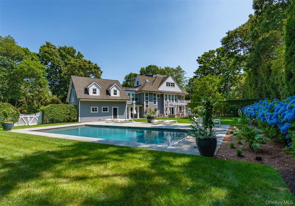 A large gray house with a gabled roof and multiple windows, surrounded by lush green trees. In front of the house, there is a rectangular swimming pool with a paved patio area. Potted plants and blooming flowers are around the pool, and the scene appears to be on a bright, sunny day in a well-maintained yard.