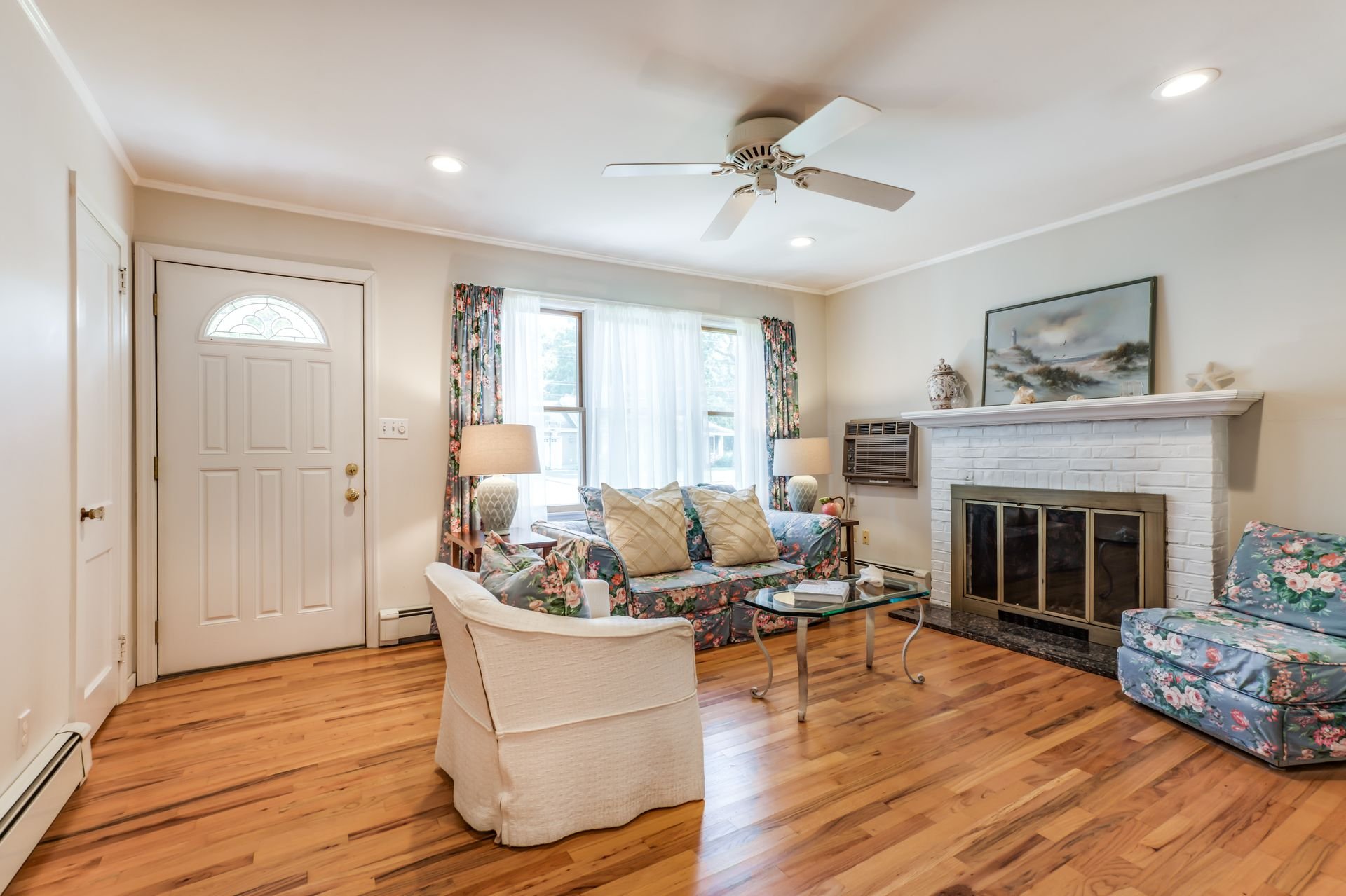 A cozy living room with hardwood floors, a white brick fireplace, and floral-patterned furniture including a sofa and chairs. Two table lamps sit on side tables flanking the window, which is dressed with floral curtains. The room features a white cei