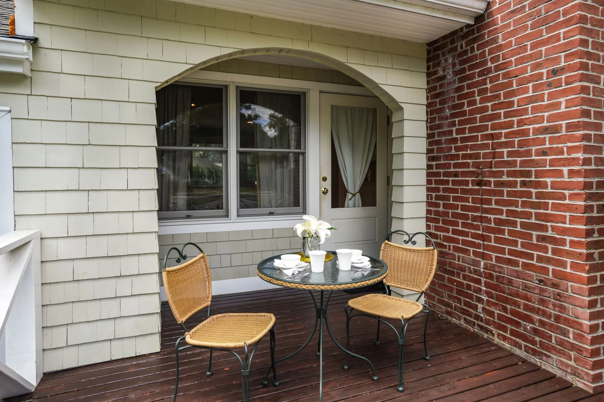 An outdoor patio with a round glass table set with white dishes and cups, surrounded by wicker chairs, against a backdrop of cream-colored and red brick walls with a window and door.