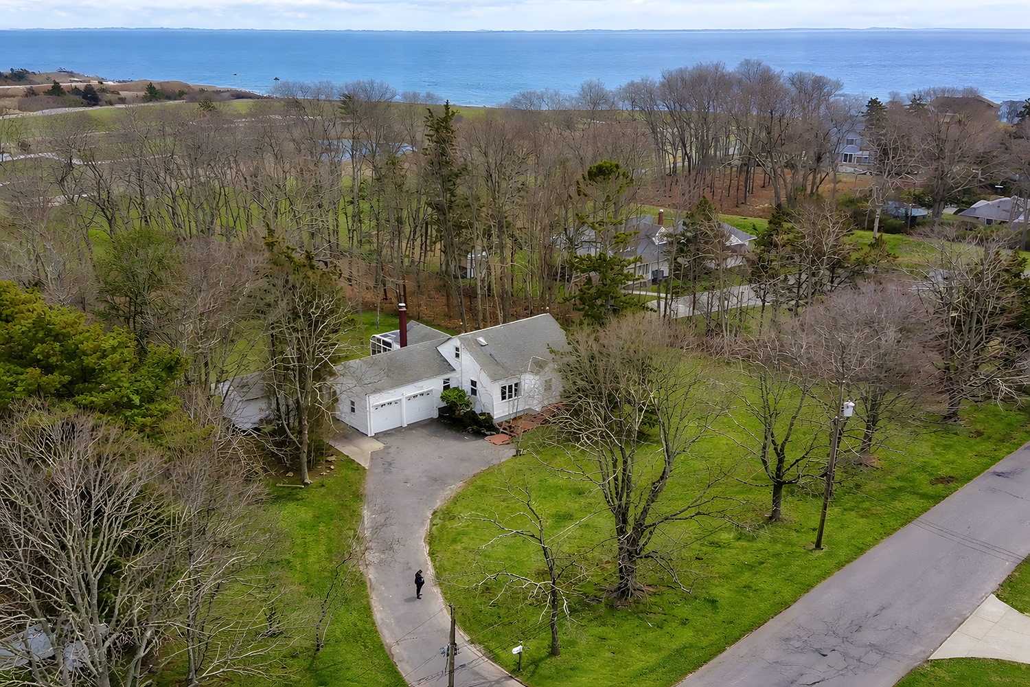 Aerial view of a white house with a driveway surrounded by leafless trees, green grass, and a distant view of the ocean under a cloudy sky.