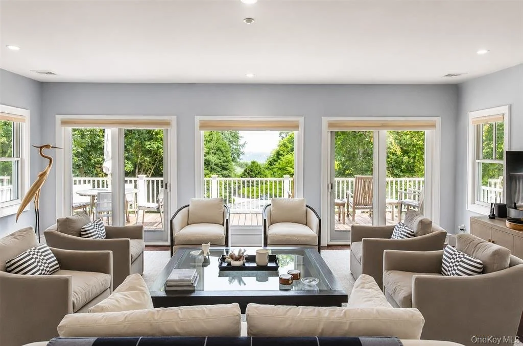 Living room with beige sofas and chairs, black coffee table, and large sliding glass doors leading to a deck with outdoor furniture and green trees outside.