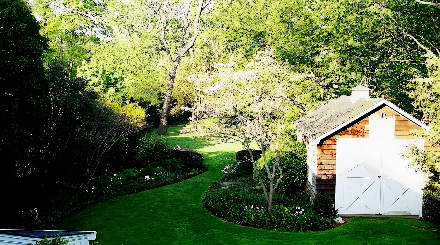 A lush green backyard with a winding grass pathway, surrounded by trees, bushes, and flowers. There is a small white shed with a shingled roof on the right side.