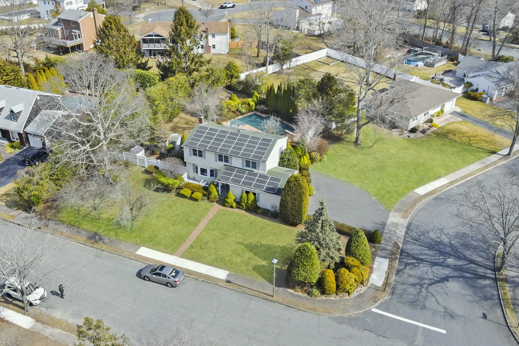 An aerial view of a neighborhood with houses, trees, lawns, and a swimming pool. One house has solar panels on the roof, and there are cars parked on the street.