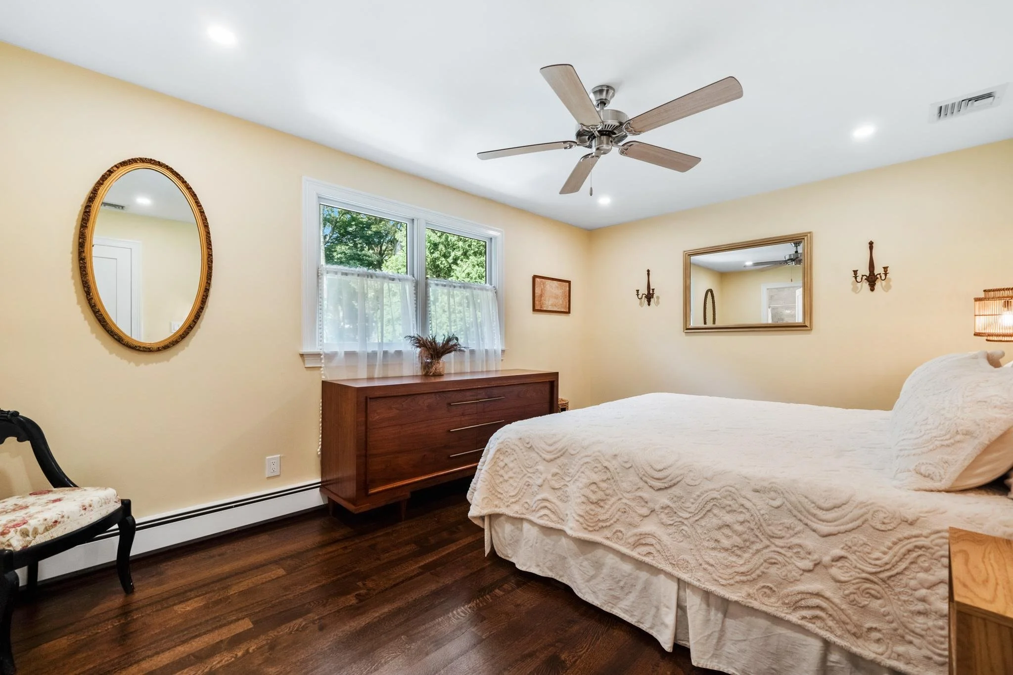 A bedroom with a ceiling fan, two windows with sheer curtains, a wooden dresser, a mirror, and a bed with a cream-colored bedspread.