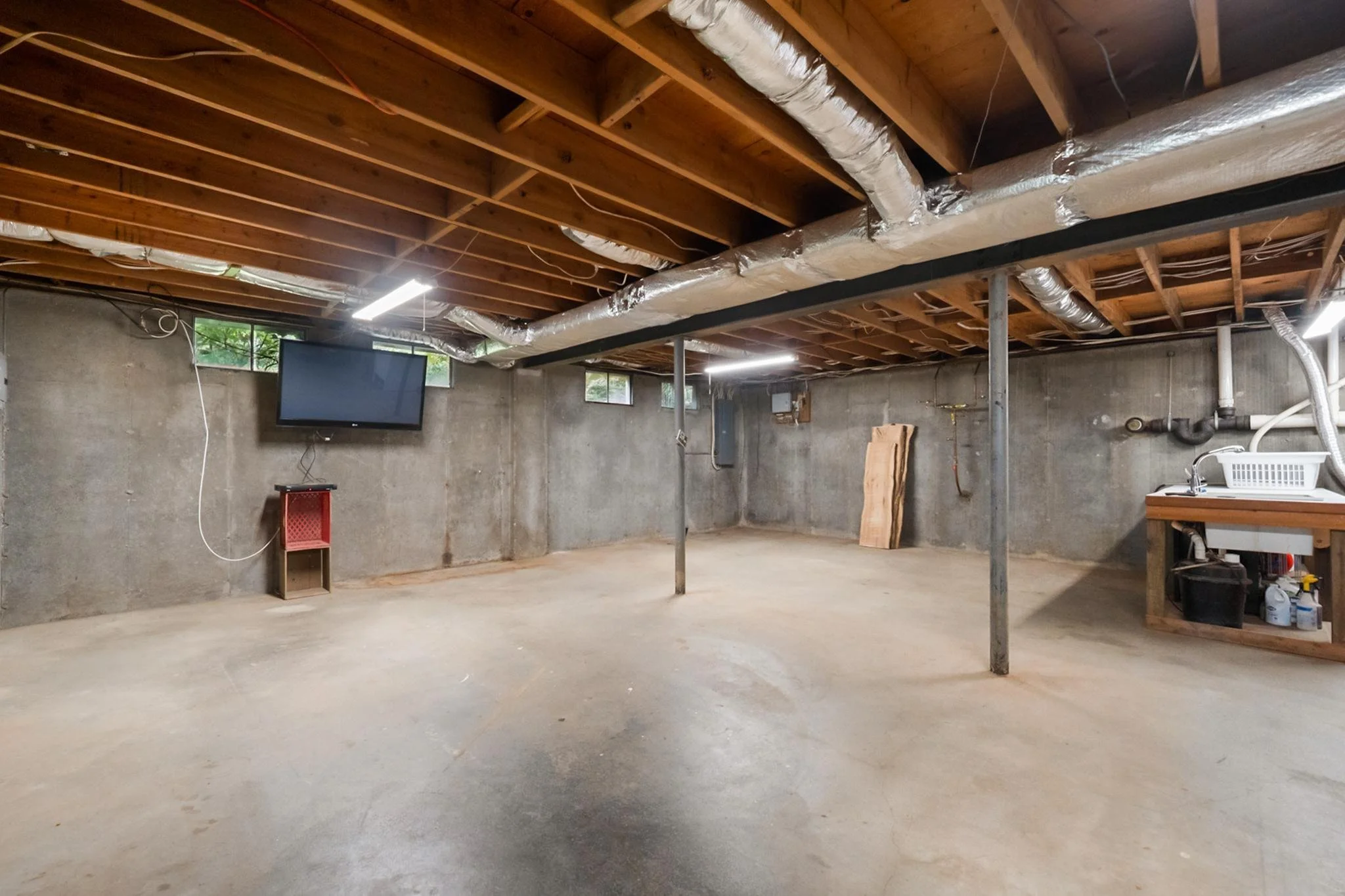 Unfinished basement with exposed wooden ceiling beams, ductwork, wall-mounted TV, and concrete walls.