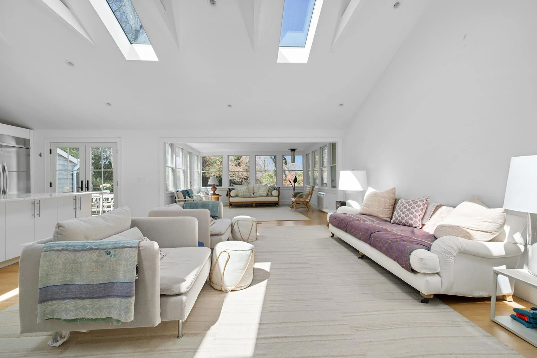 Spacious, bright living room with white walls and a vaulted ceiling with skylights, featuring multiple sofas, a coffee table, and large windows showing trees outside.