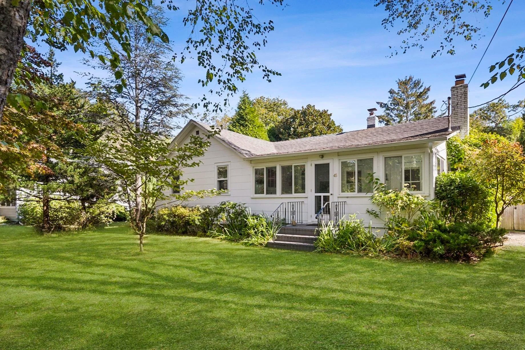 A white house with large front windows and a small front porch, surrounded by green trees and a well-maintained lawn, under a clear blue sky.