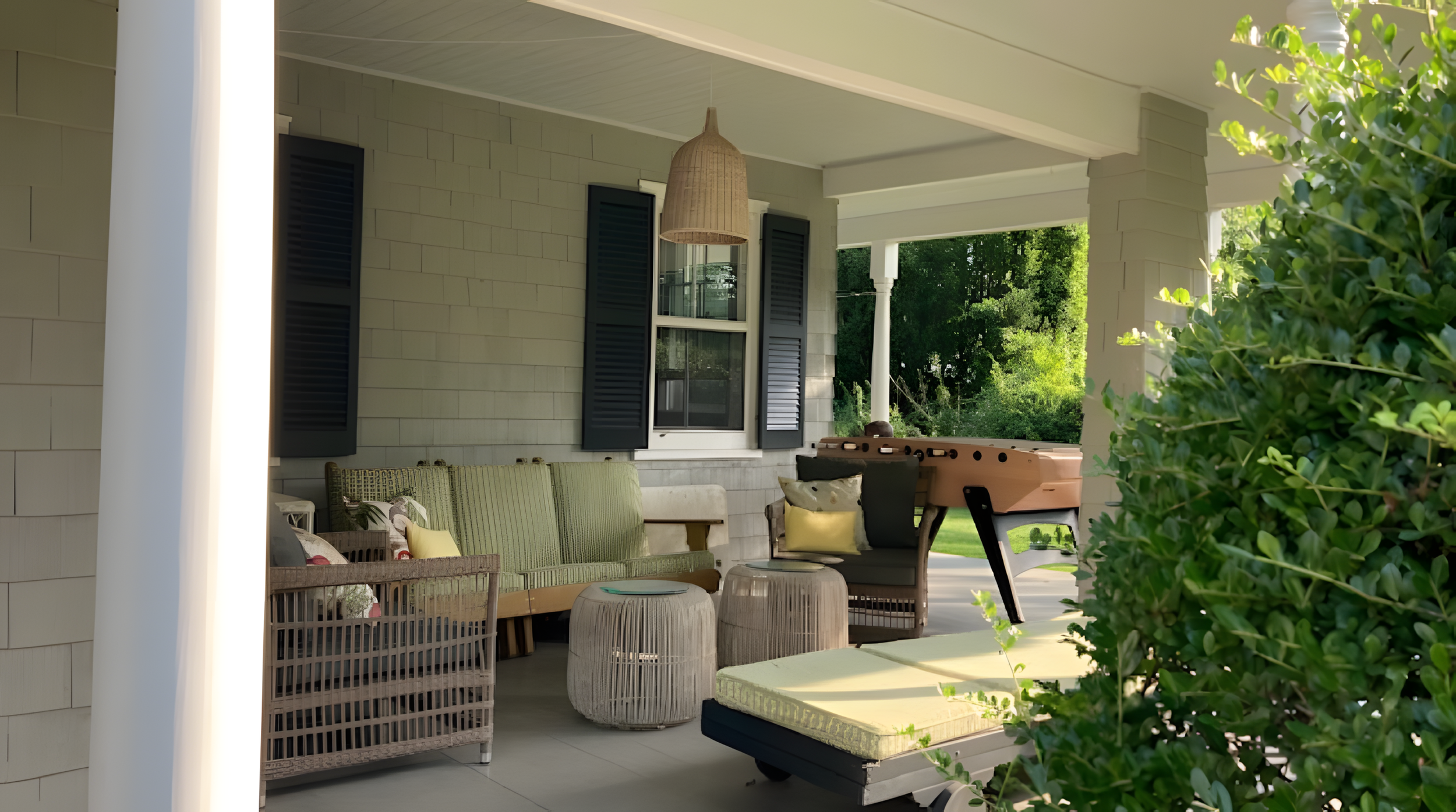 Covered porch with wicker furniture, cushions, a foosball table, and greenery in the background.