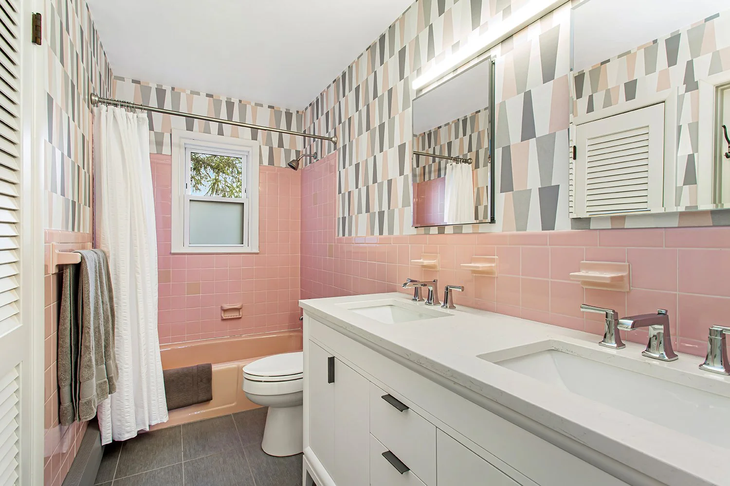 A pink-tiled bathroom with a double sink vanity, a mirror, a window with frosted glass, a bathtub with a shower, and patterned wallpaper.