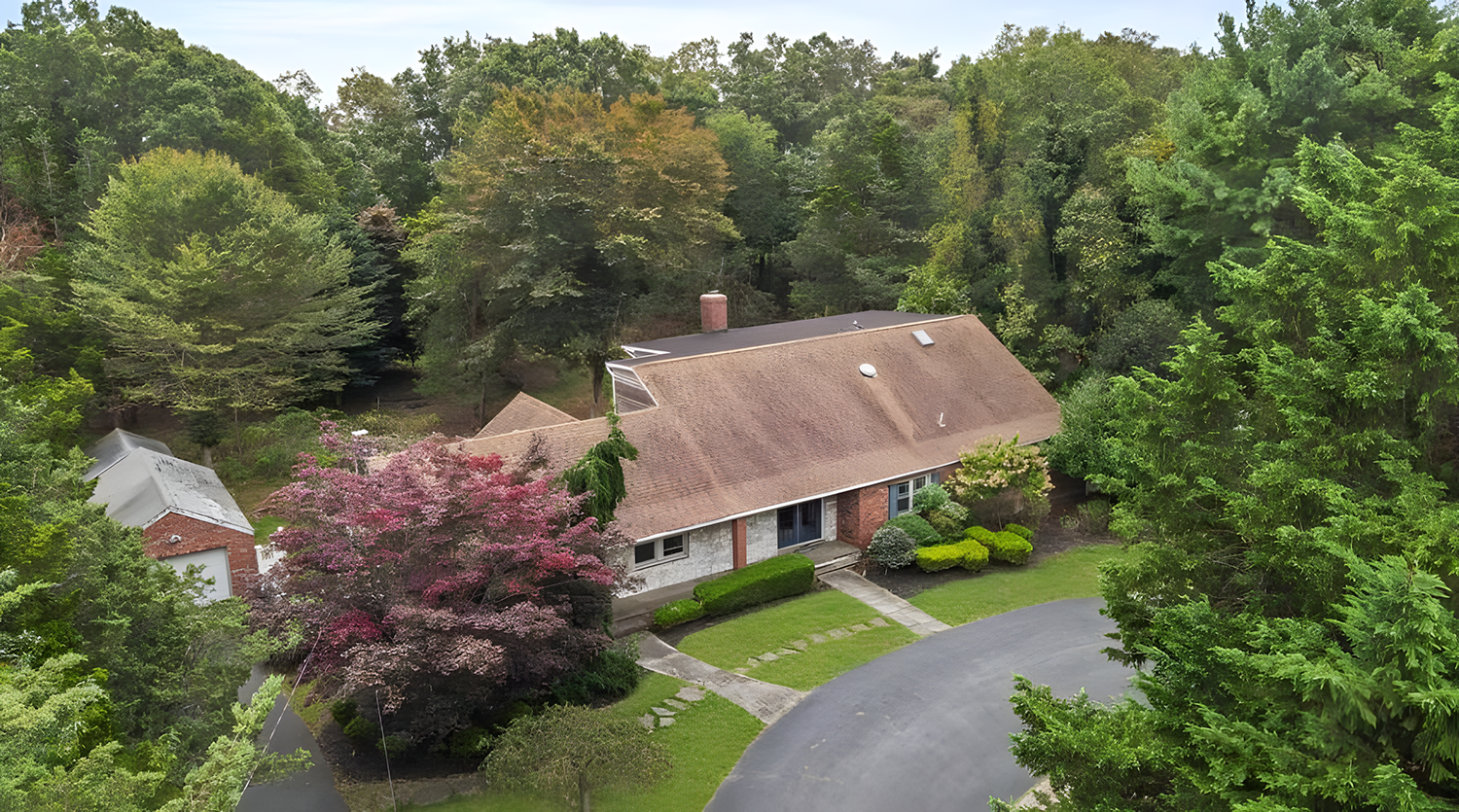 Aerial view of a single-story brick house surrounded by lush green trees and colorful flowering plants, with a curved driveway and a small front yard.