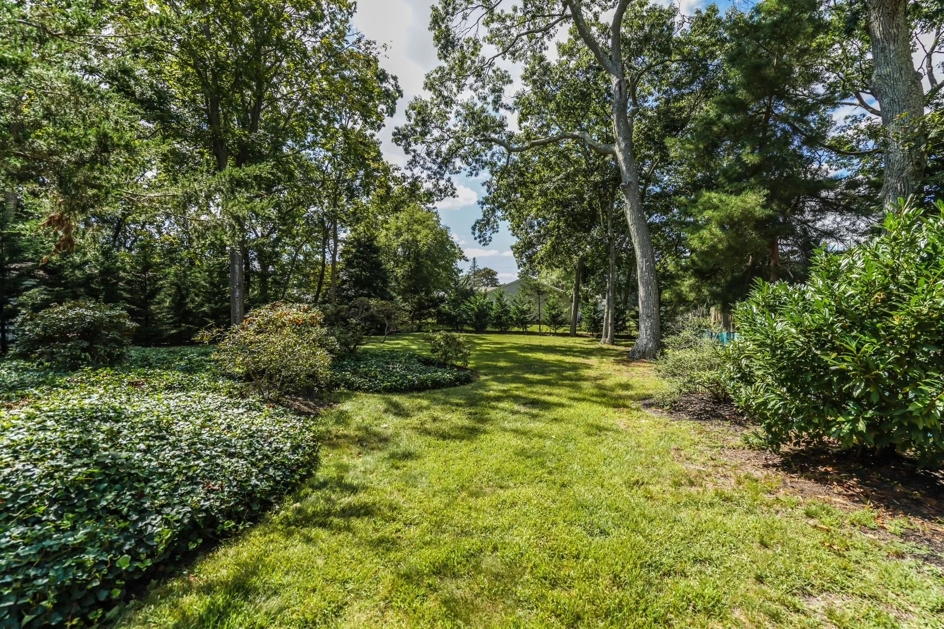 A lush backyard garden with a grassy lawn, several trees, bushes, and a hedge along the left side, under a partly cloudy sky.