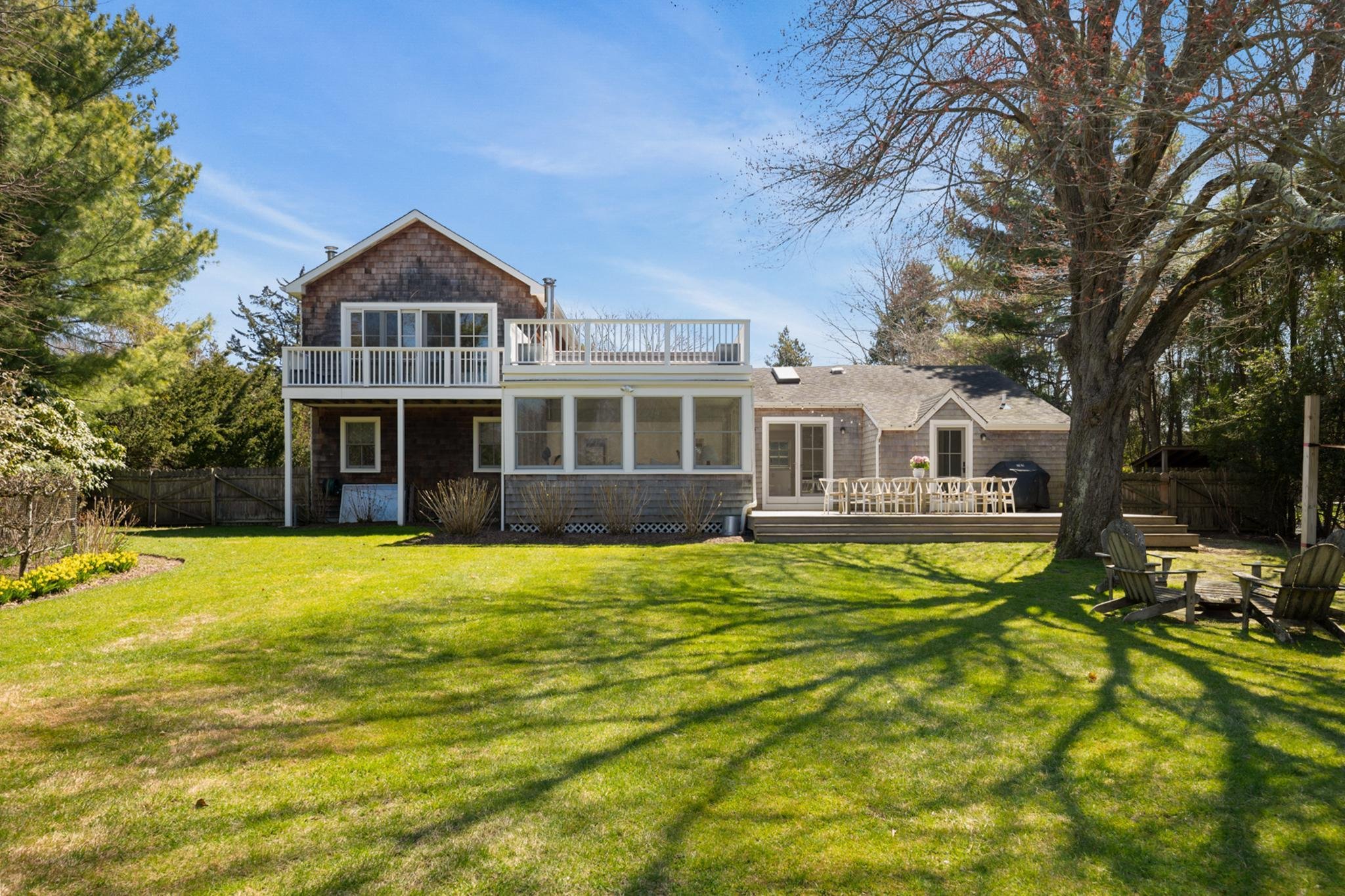 Rear view of a house with a large backyard, wooden deck, outdoor dining table, green lawn, trees, and a clear blue sky.