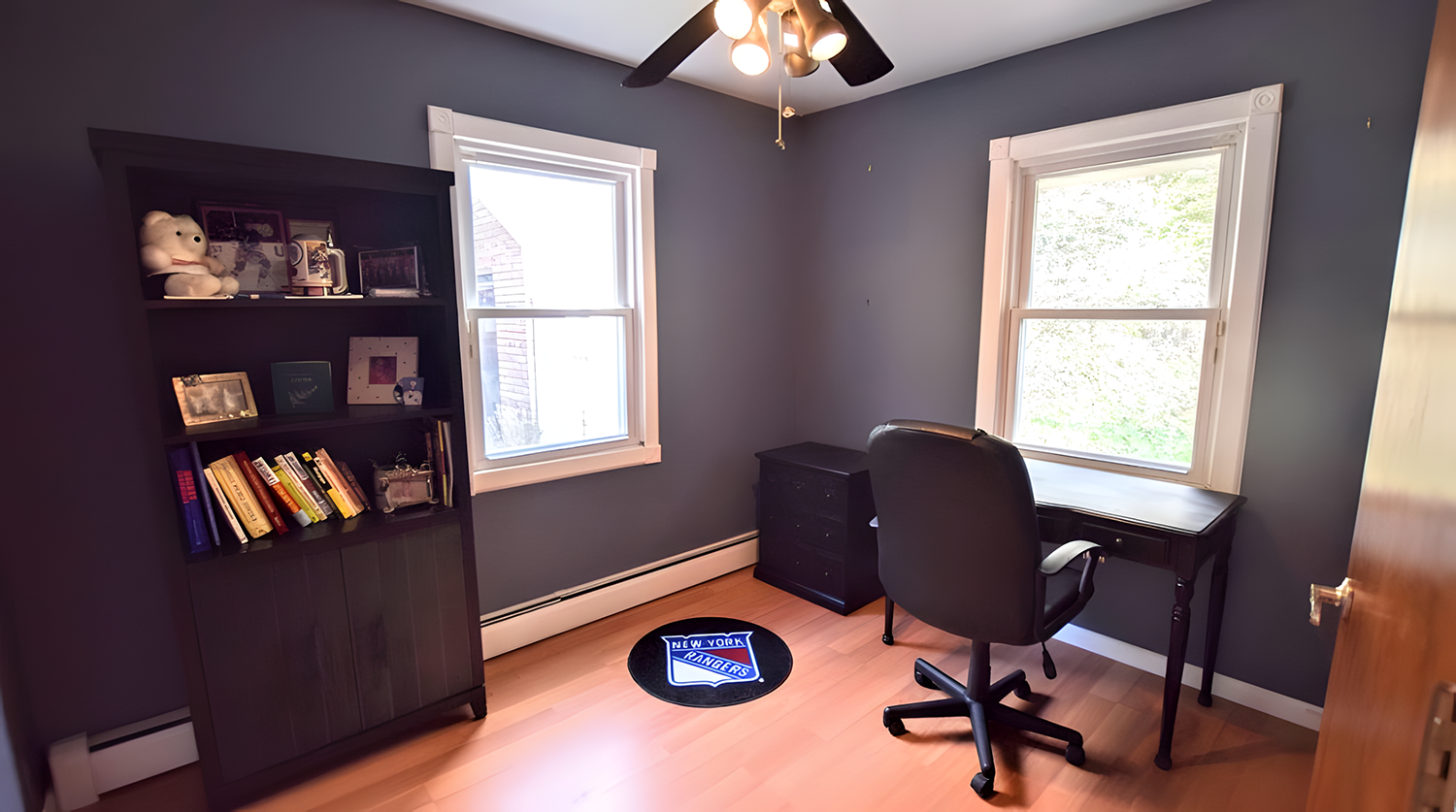 Small home office with two windows, dark wooden desk, black office chair, black bookshelf with books and stuffed animals, and a New York Rangers mat on light hardwood floor.