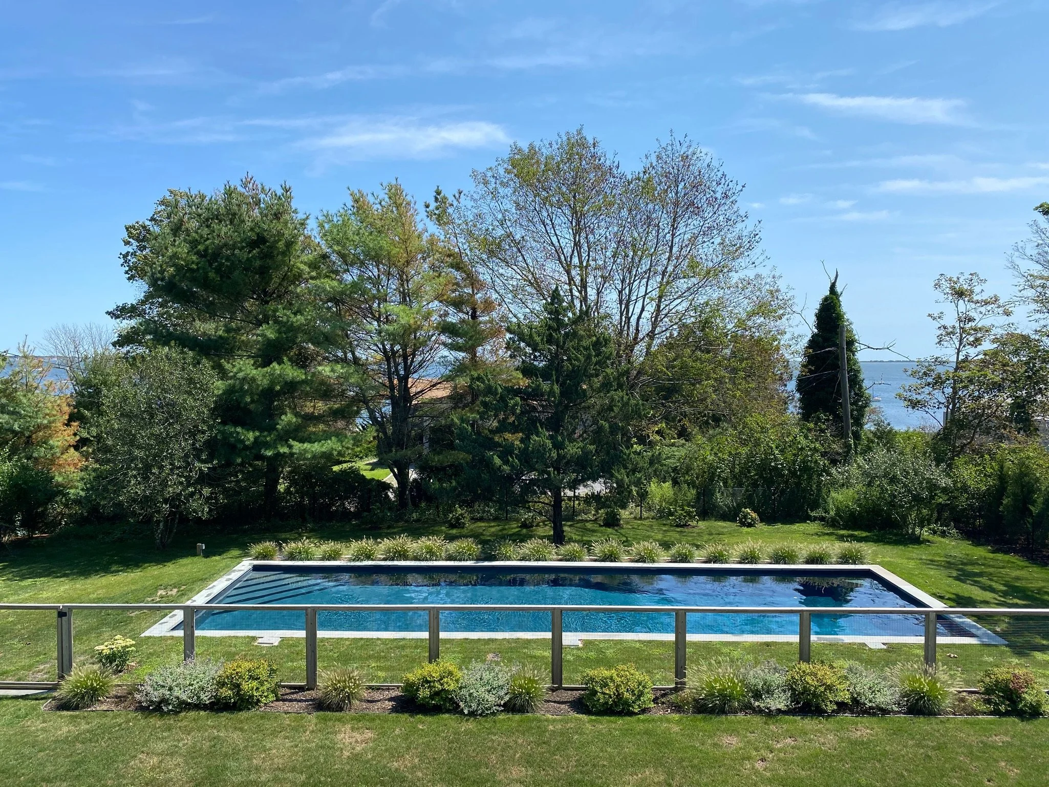A backyard with a swimming pool, green lawn, and a garden with plants, trees, and a view of water in the background under a clear blue sky.
