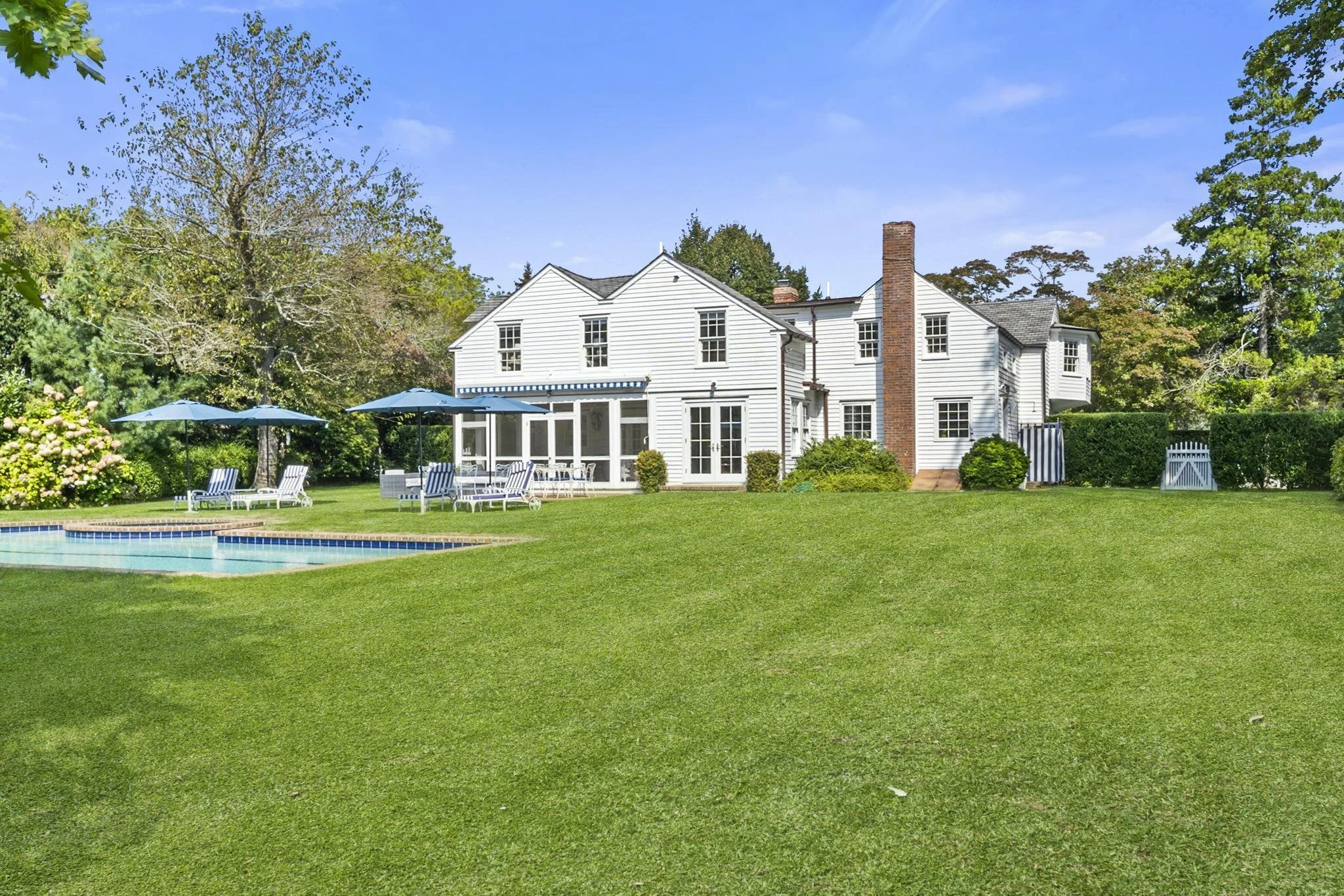 A white two-story house with a brick chimney, surrounded by a lush green lawn, trees, and bushes, with a swimming pool and lounge chairs with umbrellas in the backyard under a clear blue sky.
