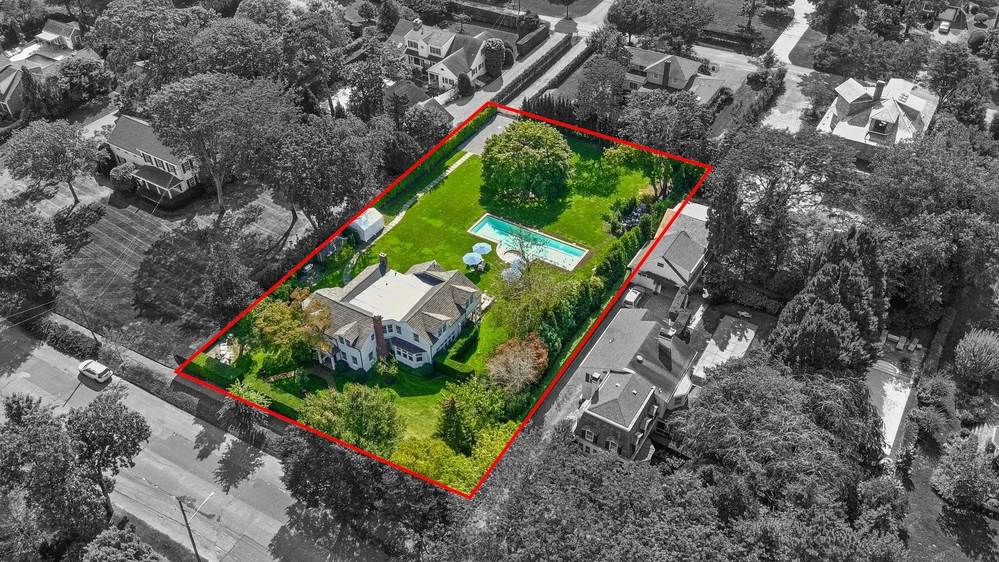 Aerial view of a residential backyard with a swimming pool, covered seating area, green lawn, surrounded by trees, with neighboring houses and streets visible.