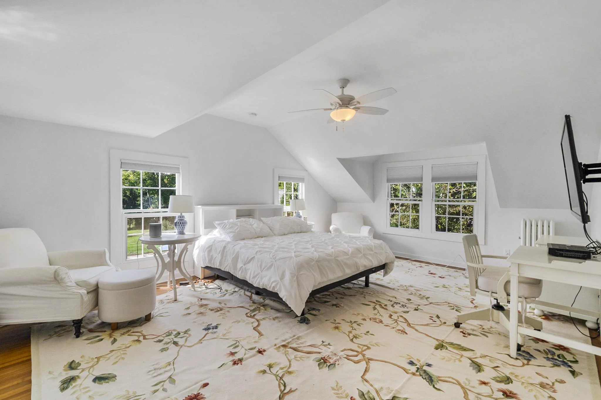 Bright bedroom with white walls, ceiling fan, and three windows overlooking greenery. Contains a white bed with bedding, a floral area rug, white armchairs, a small round white table with lamps, and a wall-mounted flat-screen TV.