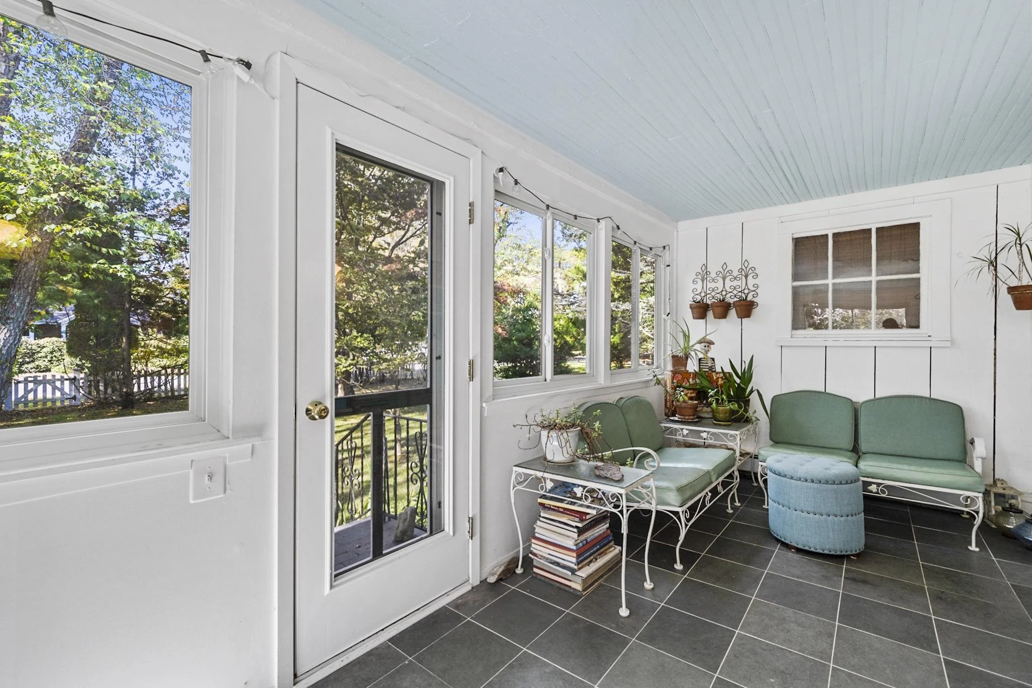 Sunroom with large windows and a door, white walls, light blue ceiling, black tiled floor, green seating, potted plants, books, and decorative items.